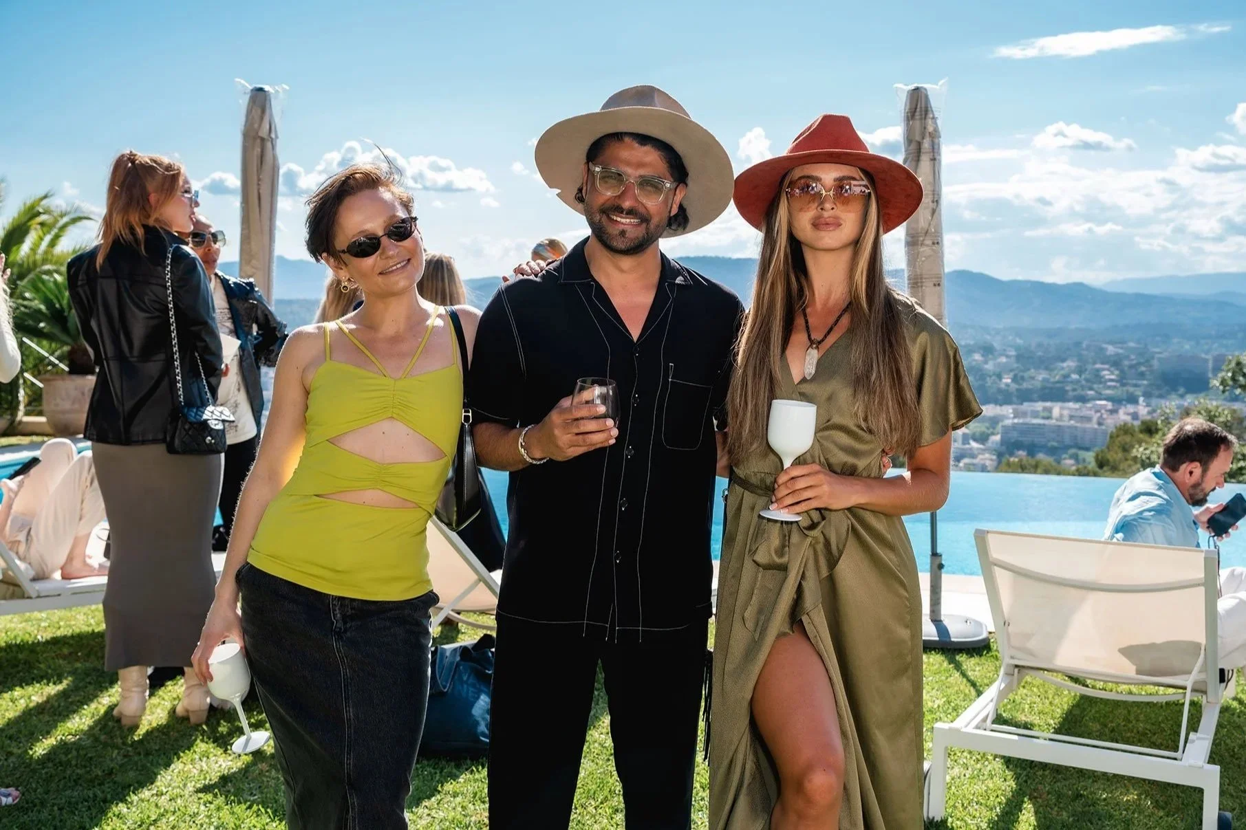 Group of three people at an outdoor gathering by a pool, with city and mountain views in the background. Two women and one man are smiling and holding wine glasses, dressed in summer attire with wide-brimmed hats and sunglasses. Others are socializin