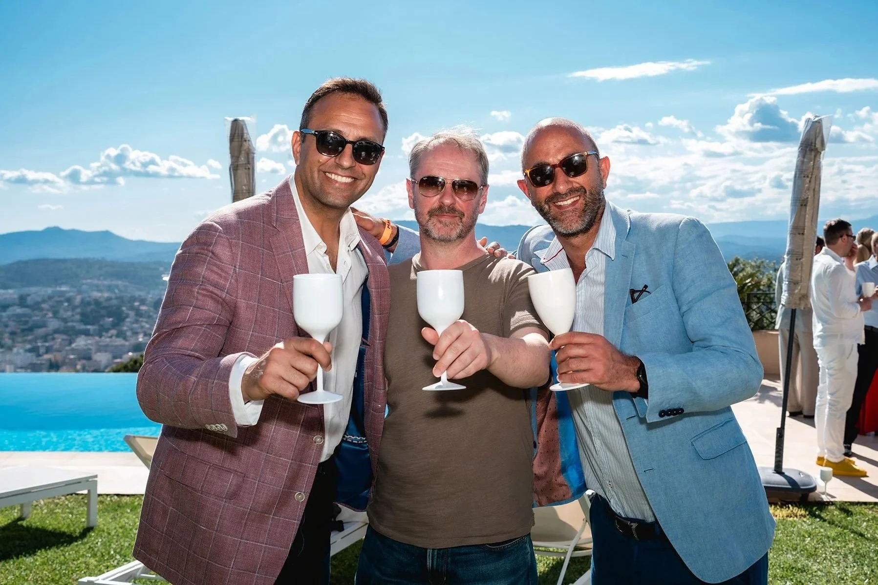 Three men wearing sunglasses and smiling while holding white wine glasses at an outdoor celebration, with a mountain view and other guests in the background.