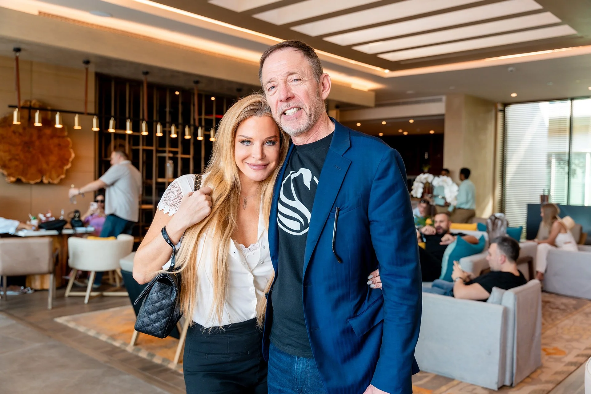 A man and woman standing close together, smiling at the camera in a hotel lobby or lounge area with other people in the background.