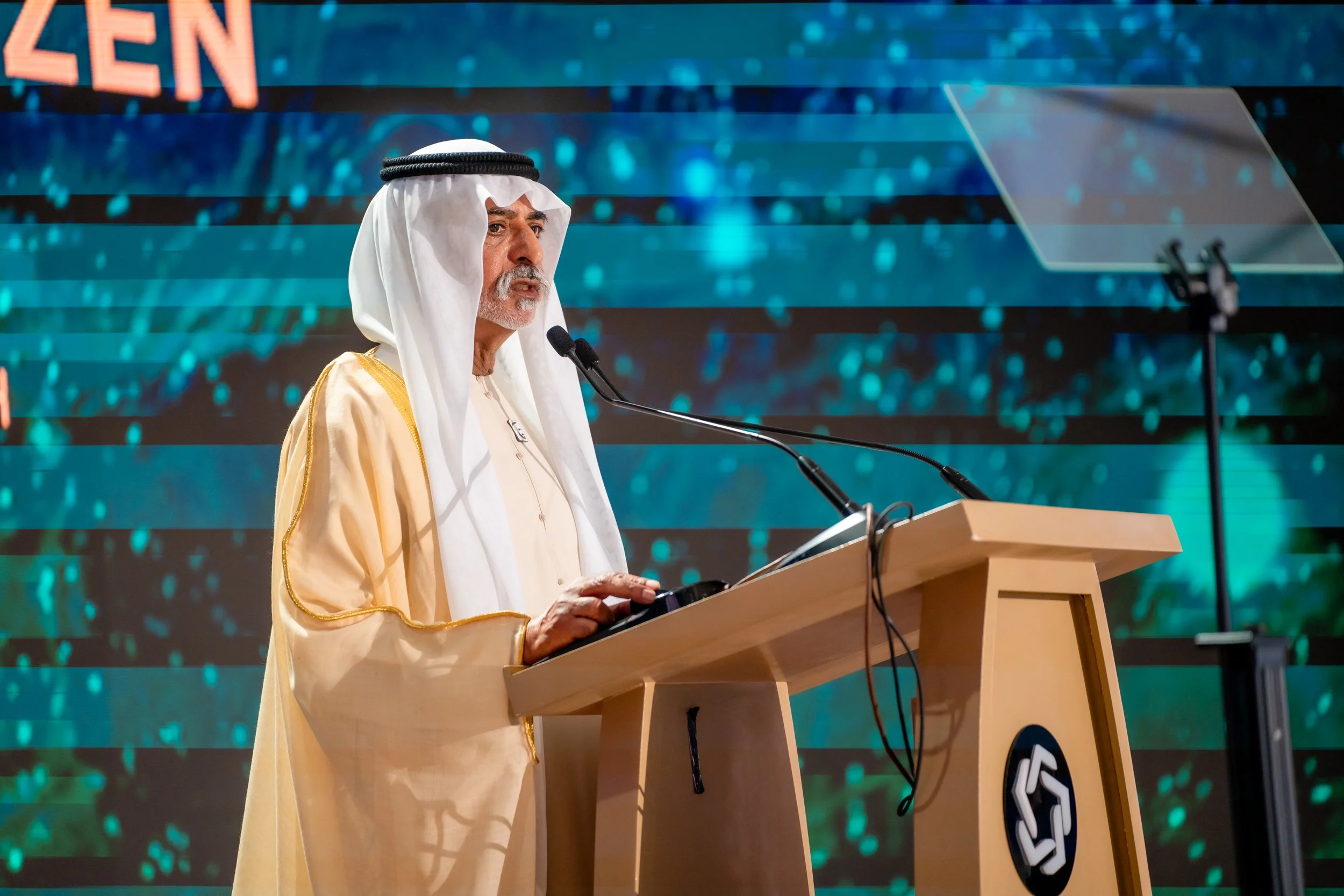 An elderly man in traditional Middle Eastern attire, including a white thobe and a head covering, stands at a wooden podium speaking into microphones during a conference or event. The background features a digital blue and teal patterned screen with 