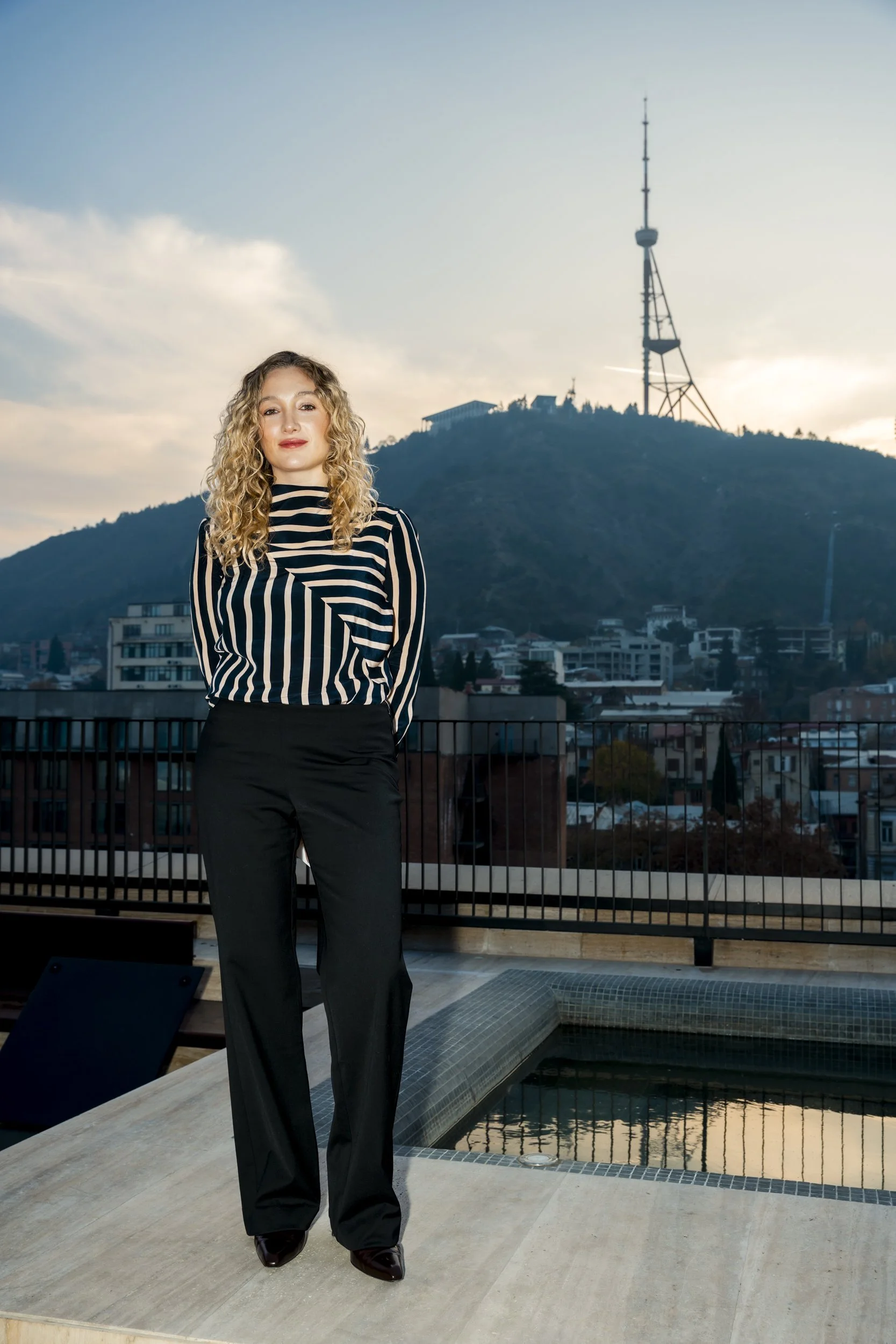 A woman with curly blonde hair standing on a balcony next to a small pool, overlooking a city with a hill and a large antenna tower in the background during sunset.