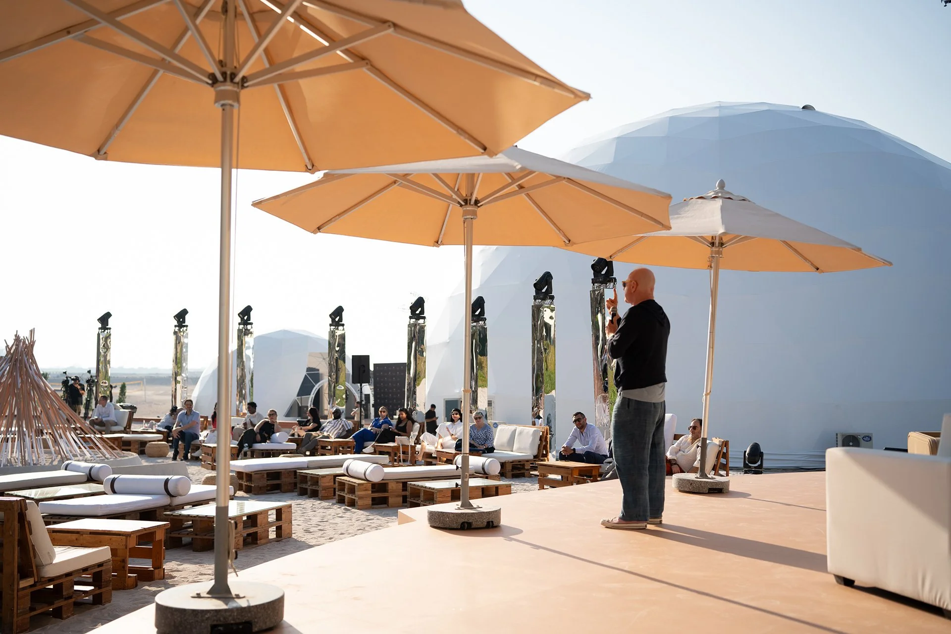 Man speaking on stage with umbrellas and lounge chairs on beach in background