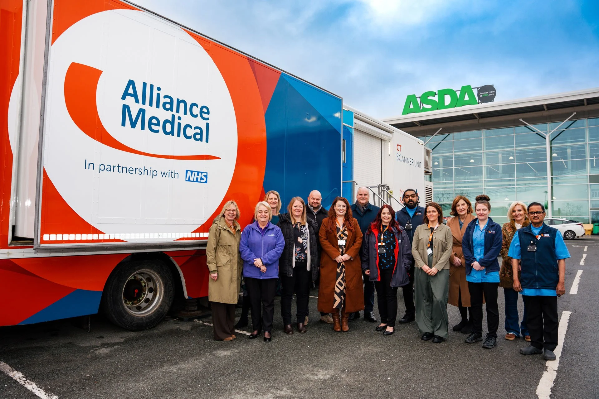 A group of healthcare workers and staff standing in front of a mobile medical unit outside an ASDA store, with a large Alliance Medical logo and NHS partnership branding on the vehicle.