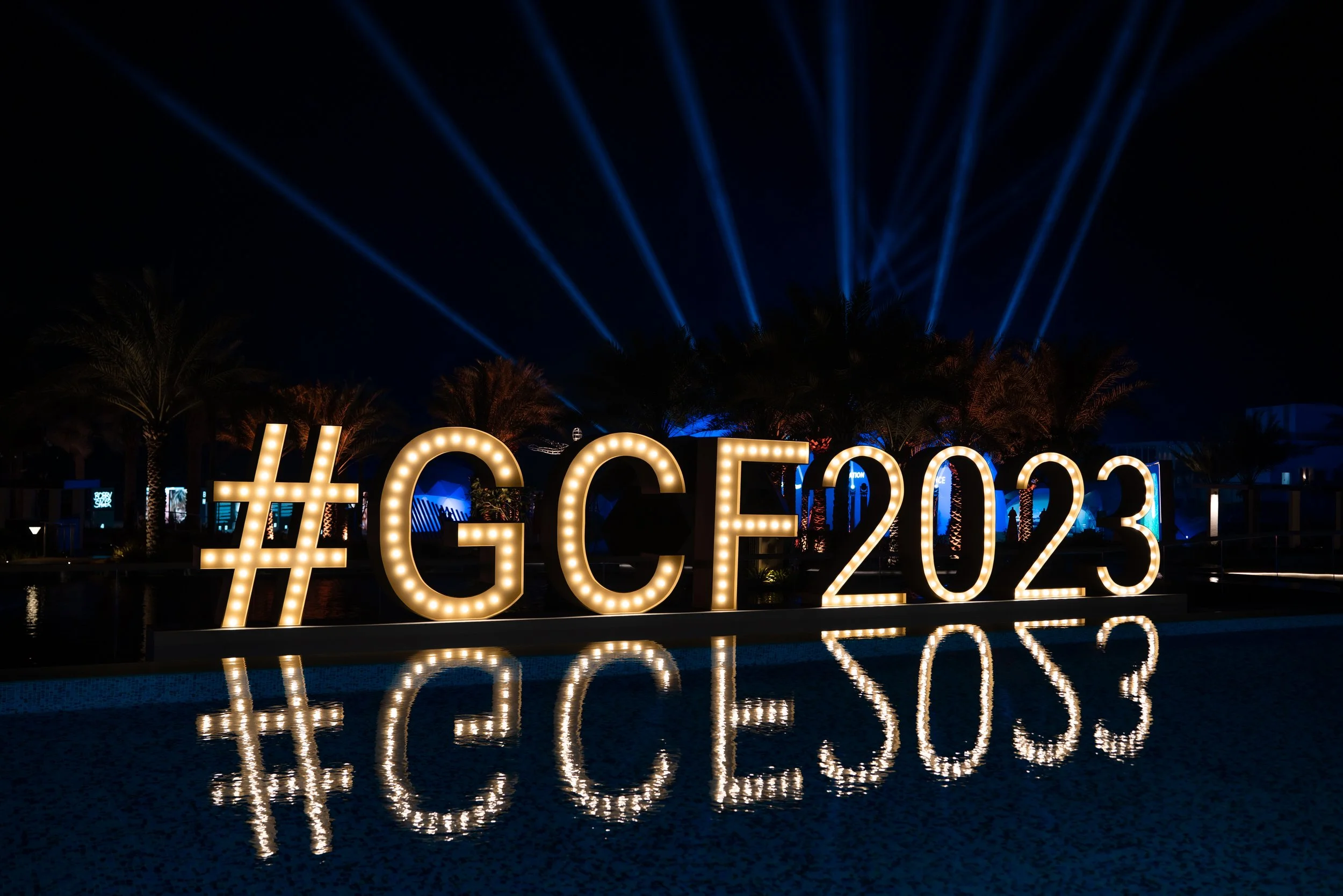 Illuminated hashtags and letters '#GCF2023' reflected on a pool at night, with palm trees and blue spotlights in the background.