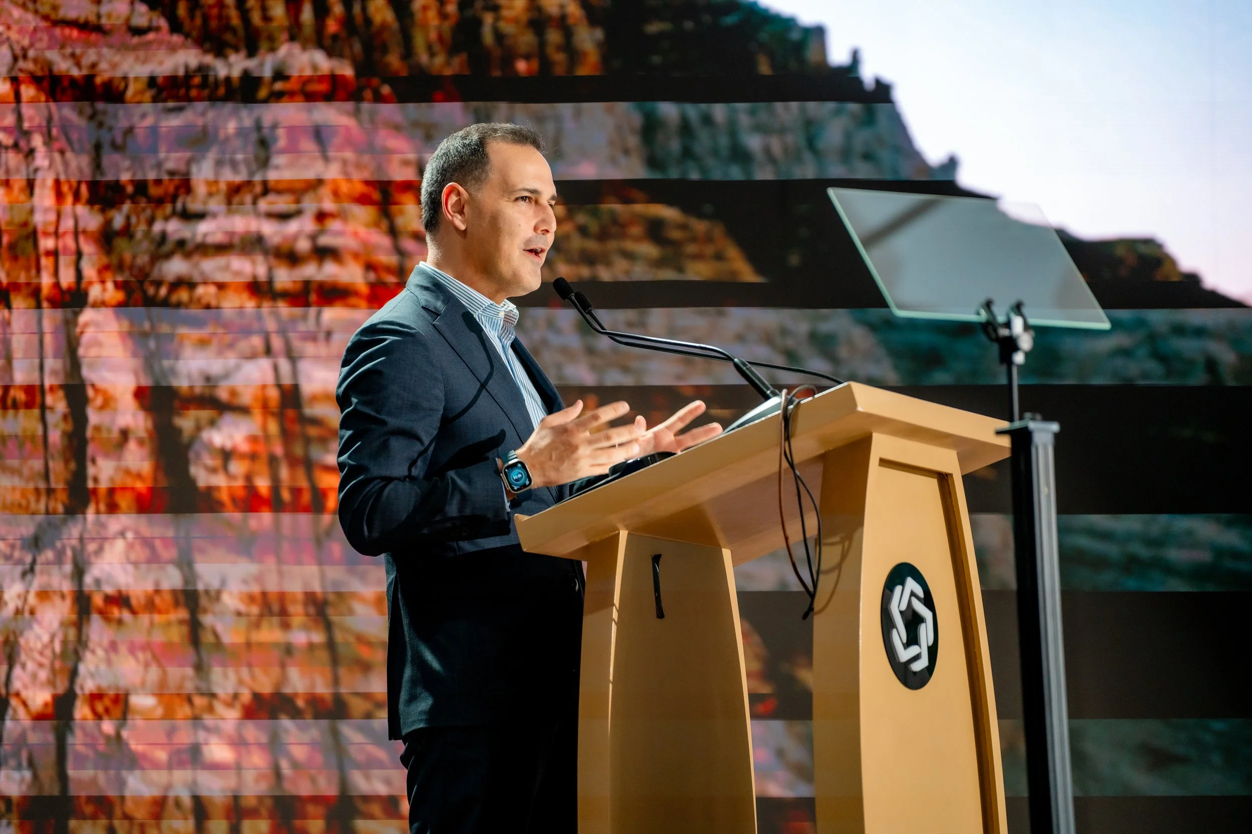 A man in a dark suit giving a presentation at a wooden podium with a logo, standing in front of a large screen displaying an abstract image of a rocky landscape with red and orange hues.