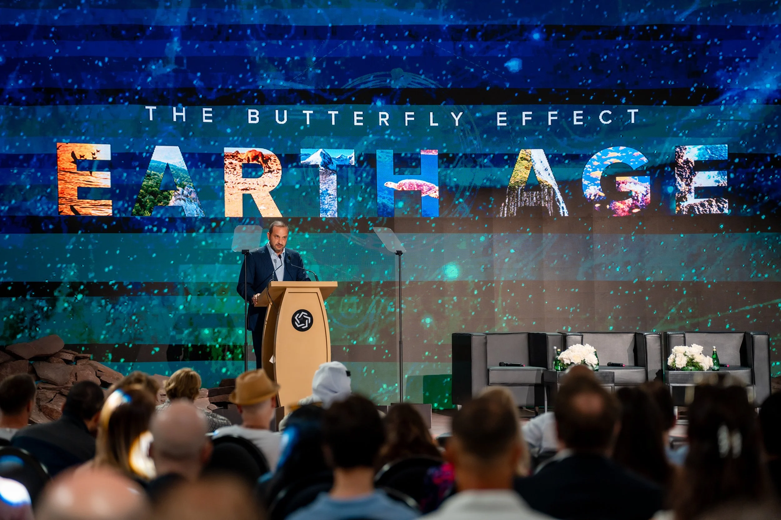 A man in a dark suit giving a presentation at a conference, standing behind a wooden podium with a logo, with a large screen behind him displaying "The Butterfly Effect" and "Earth Age" in colorful text, in front of an audience.