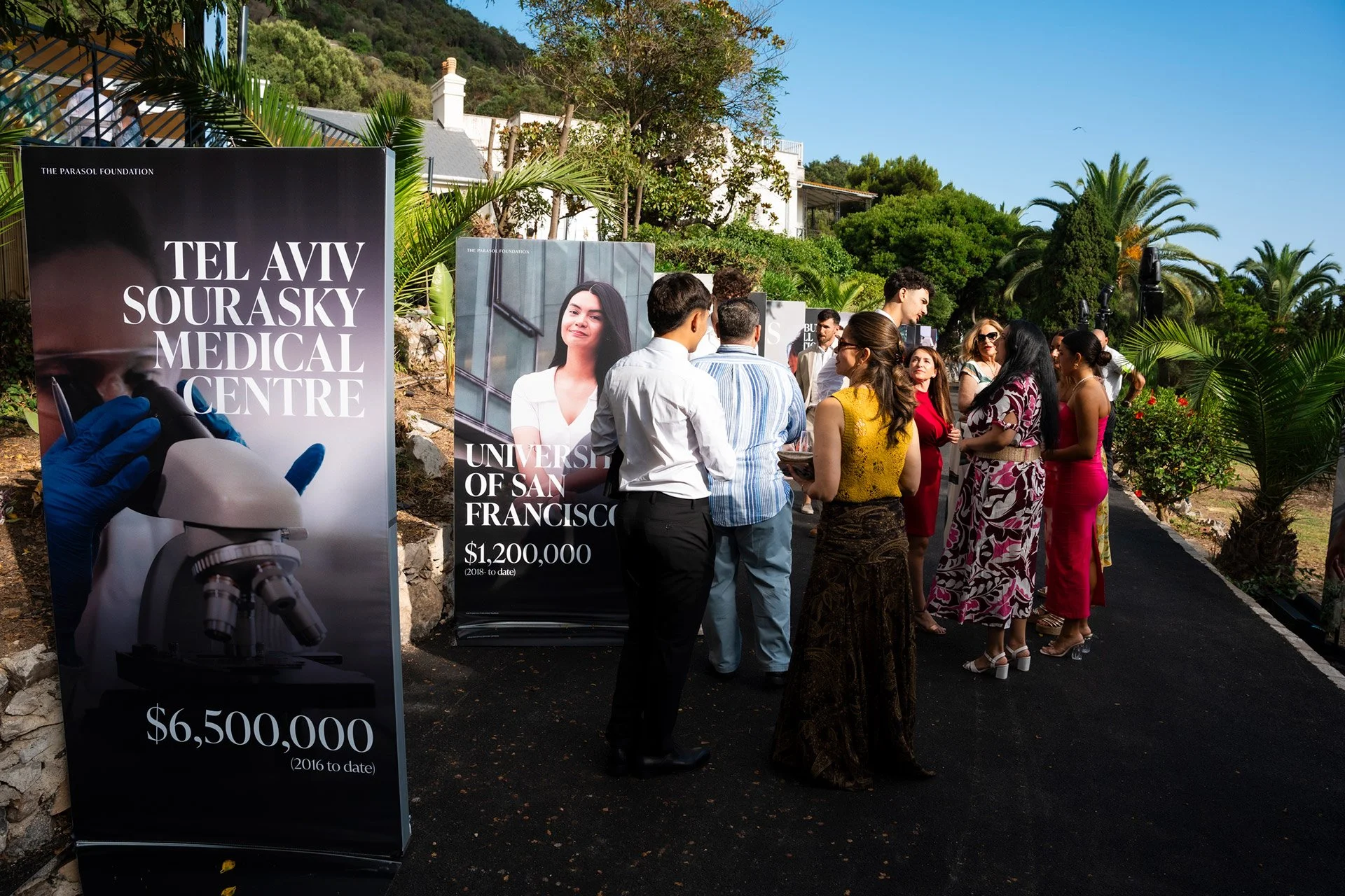 Group of people at an outdoor event near informational banners about medical and university research, with lush greenery and clear blue sky in the background.