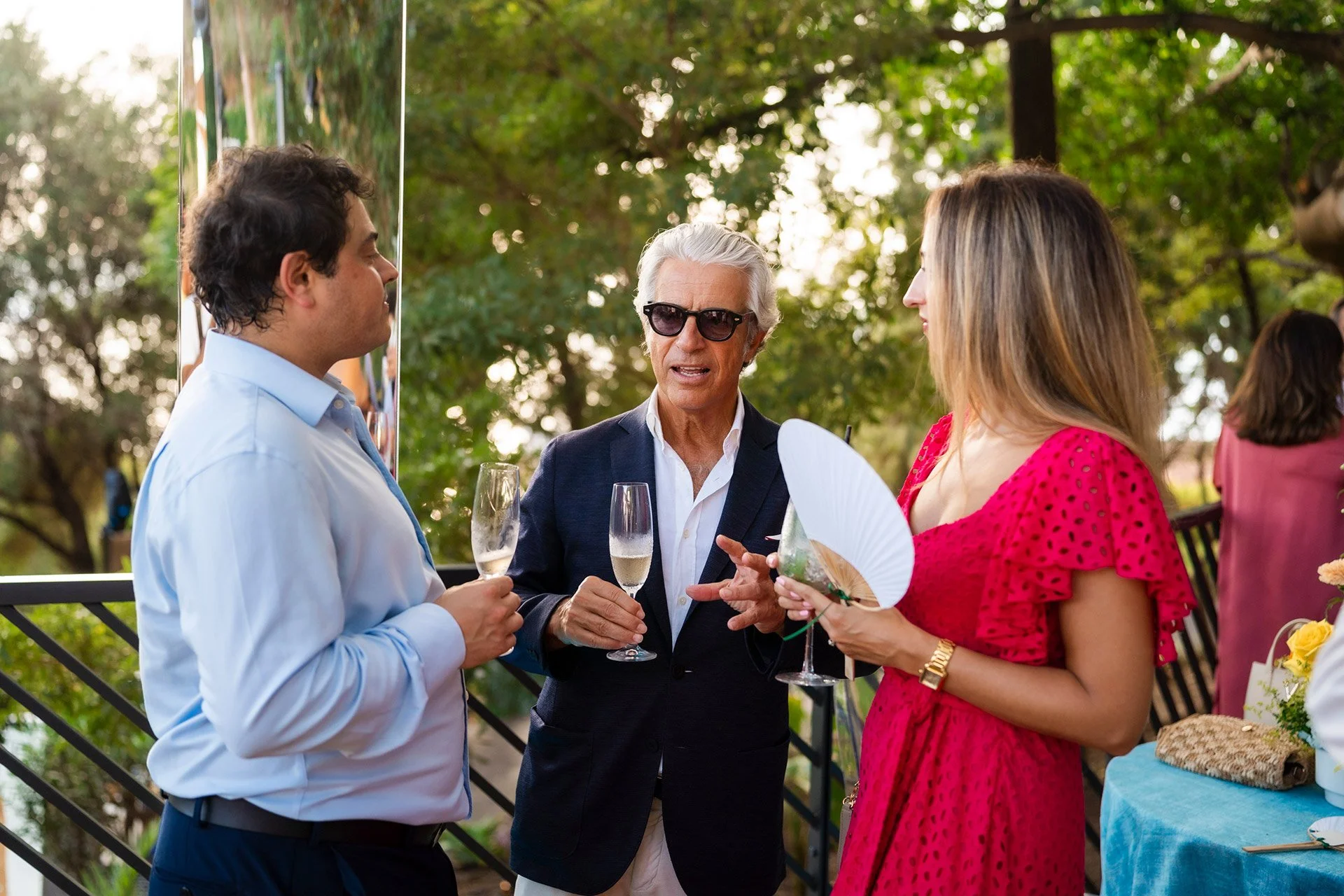 Three people having a conversation at an outdoor event, holding champagne glasses.