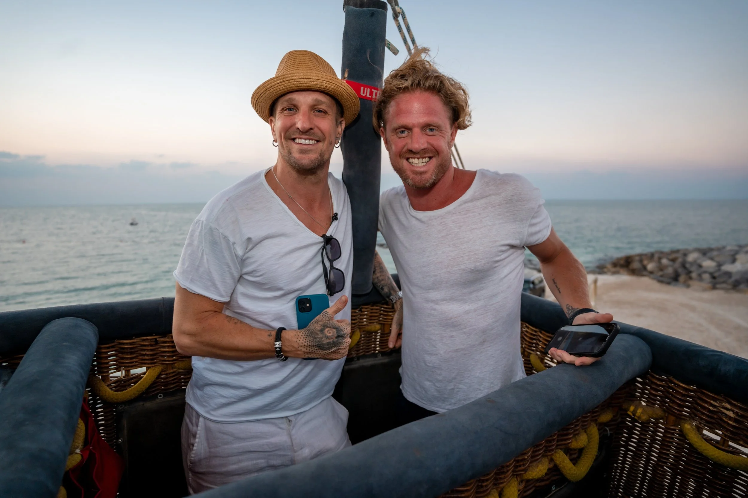 Two men smiling inside a hot air balloon basket near a body of water at sunset.