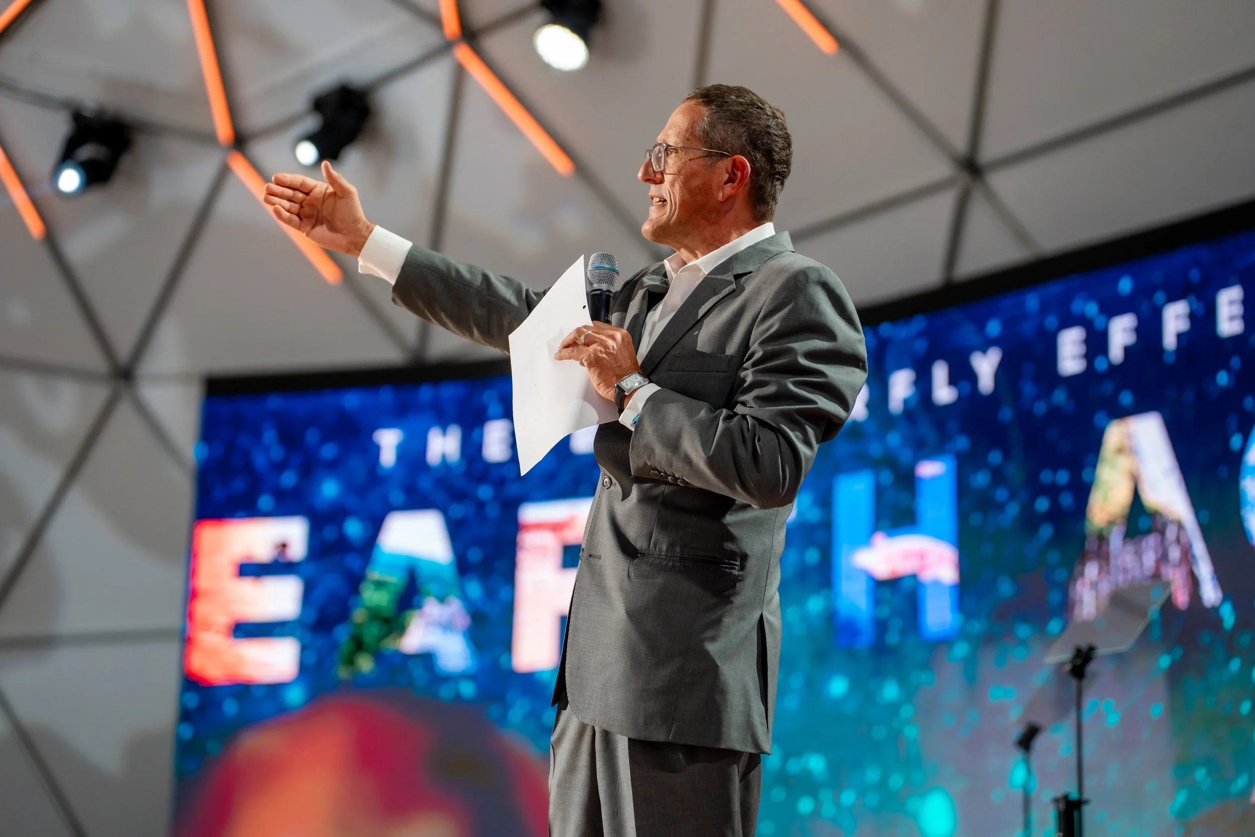 A man in a gray suit and glasses is speaking with a microphone in his right hand and a piece of paper in his left hand. He is gesturing with his left hand, and there is a large digital screen behind him with partially visible text and graphics.
