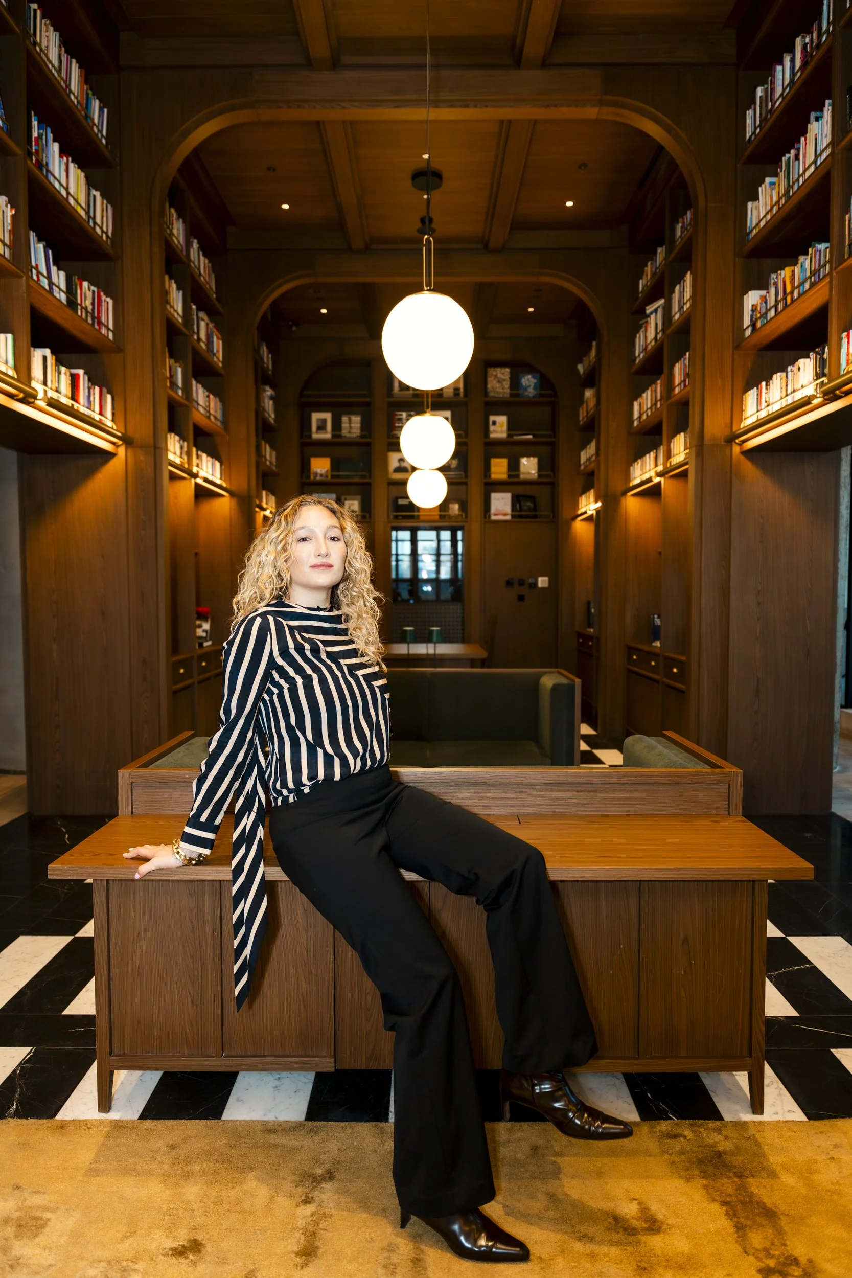 A woman with curly blonde hair dressed in a black-and-white striped top and black pants sitting on a wooden bench in a library with tall wooden bookshelves and hanging spherical lights.