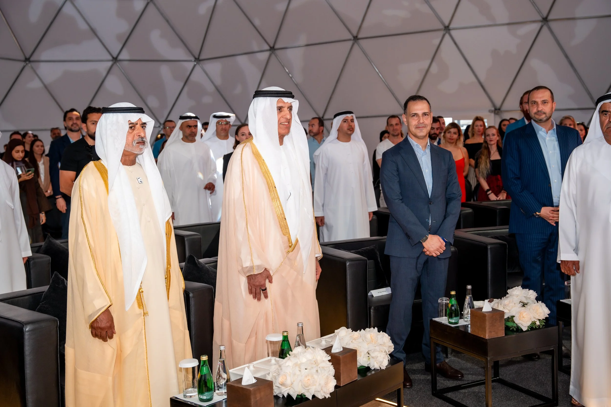 Group of men and women standing during a formal event or conference, some dressed in traditional Middle Eastern attire, in a large, dome-like indoor venue with tables and flowers in the foreground.
