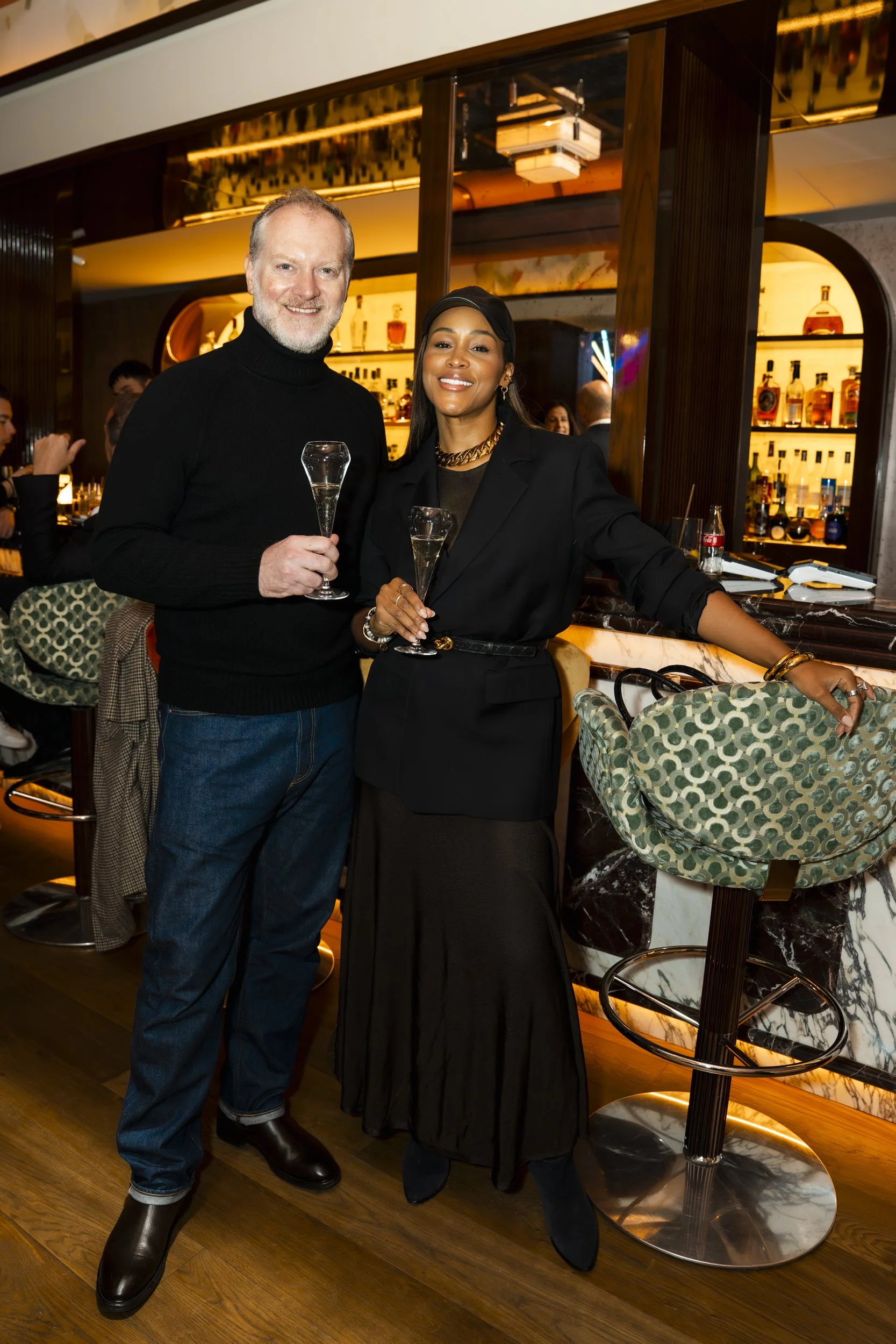 A man and a woman dressed elegantly at a bar, holding champagne glasses, with a well-lit bar and seating area in the background.