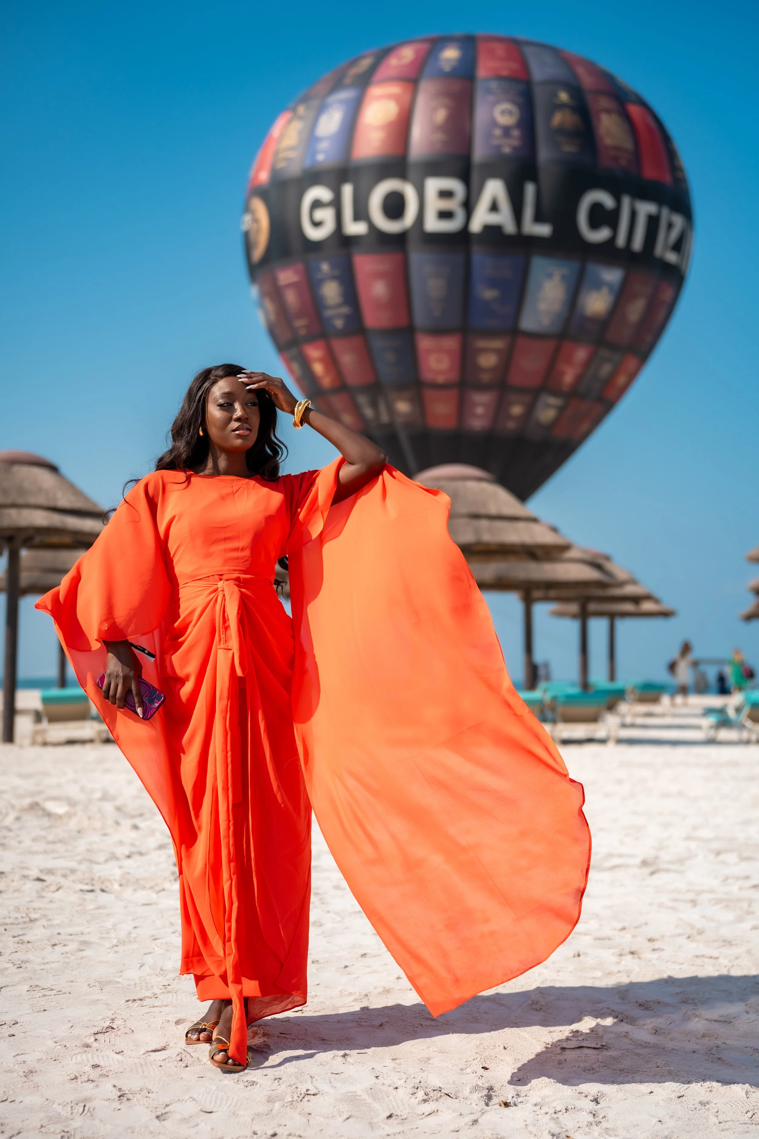 A woman in an orange dress standing on a beach with hot air balloons in the background, one of which has 'GLOBAL CITZ' written on it.
