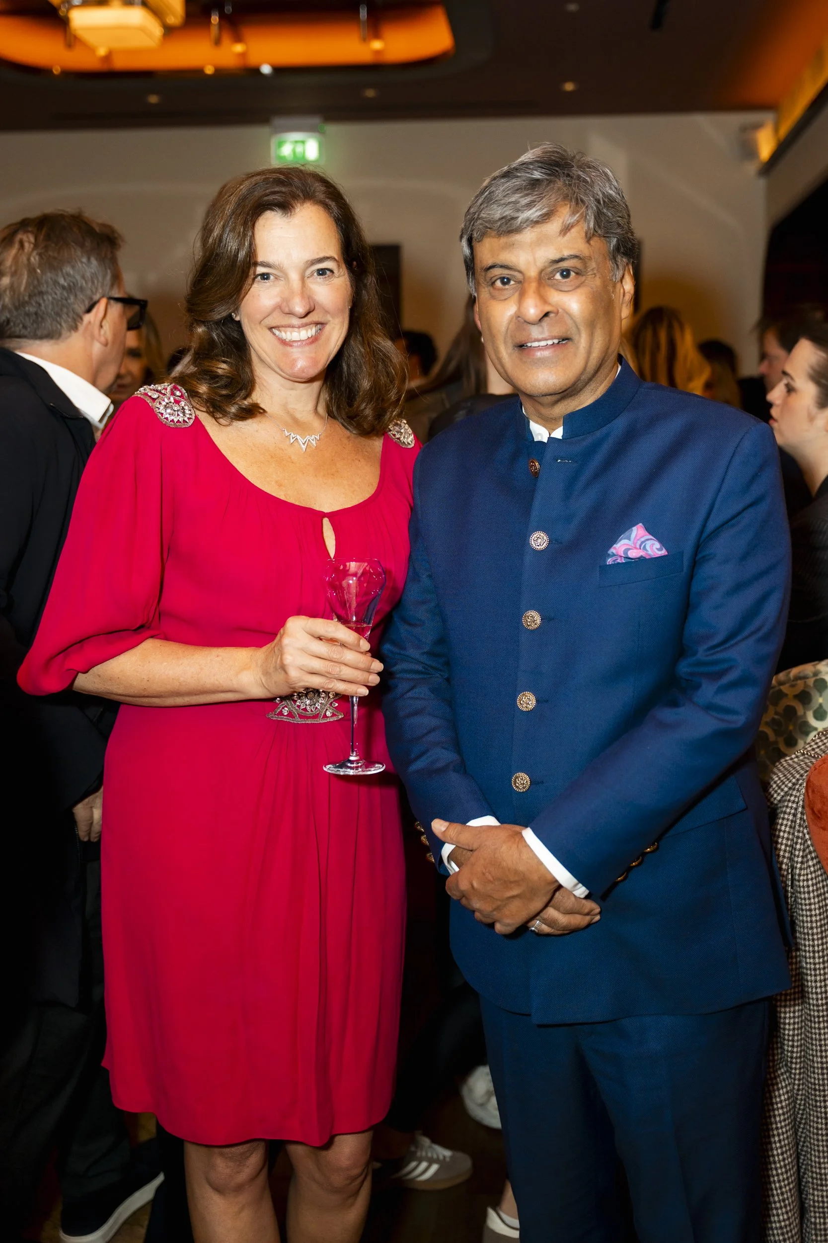 A woman in a red dress holding a glass of wine and a man in a traditional Indian blue suit at a party or formal event.