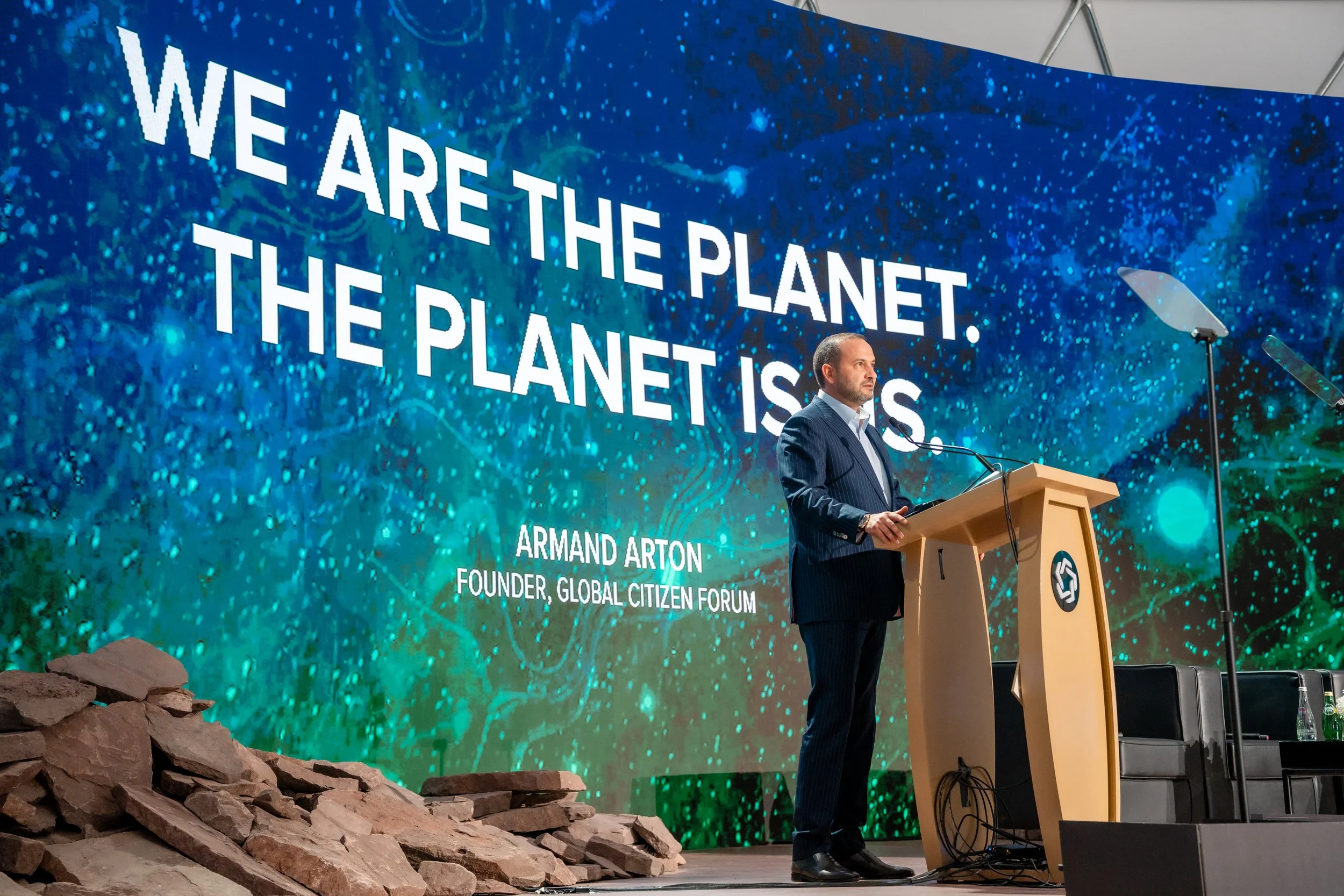 Man in a suit giving a speech at a podium on a stage with a large screen behind him displaying the text: We are the planet. The planet is us. Presented by Armand Arton, founder of Global Citizen Forum. The stage has fake rocks in the foreground.