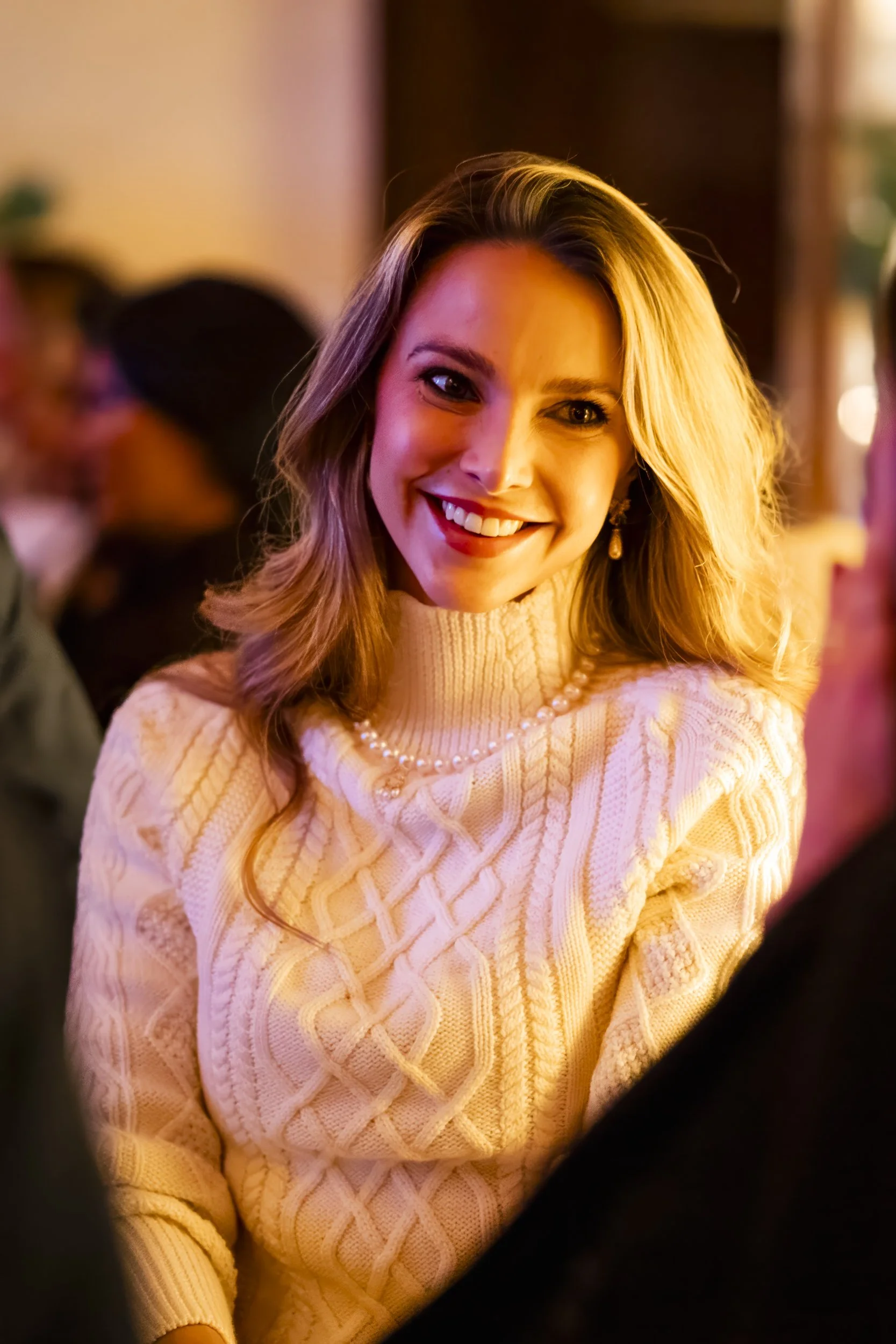 A woman with shoulder-length blonde hair, wearing a cream-colored cable knit turtleneck sweater, a pearl necklace, and earrings, smiling warmly at a social gathering indoors.