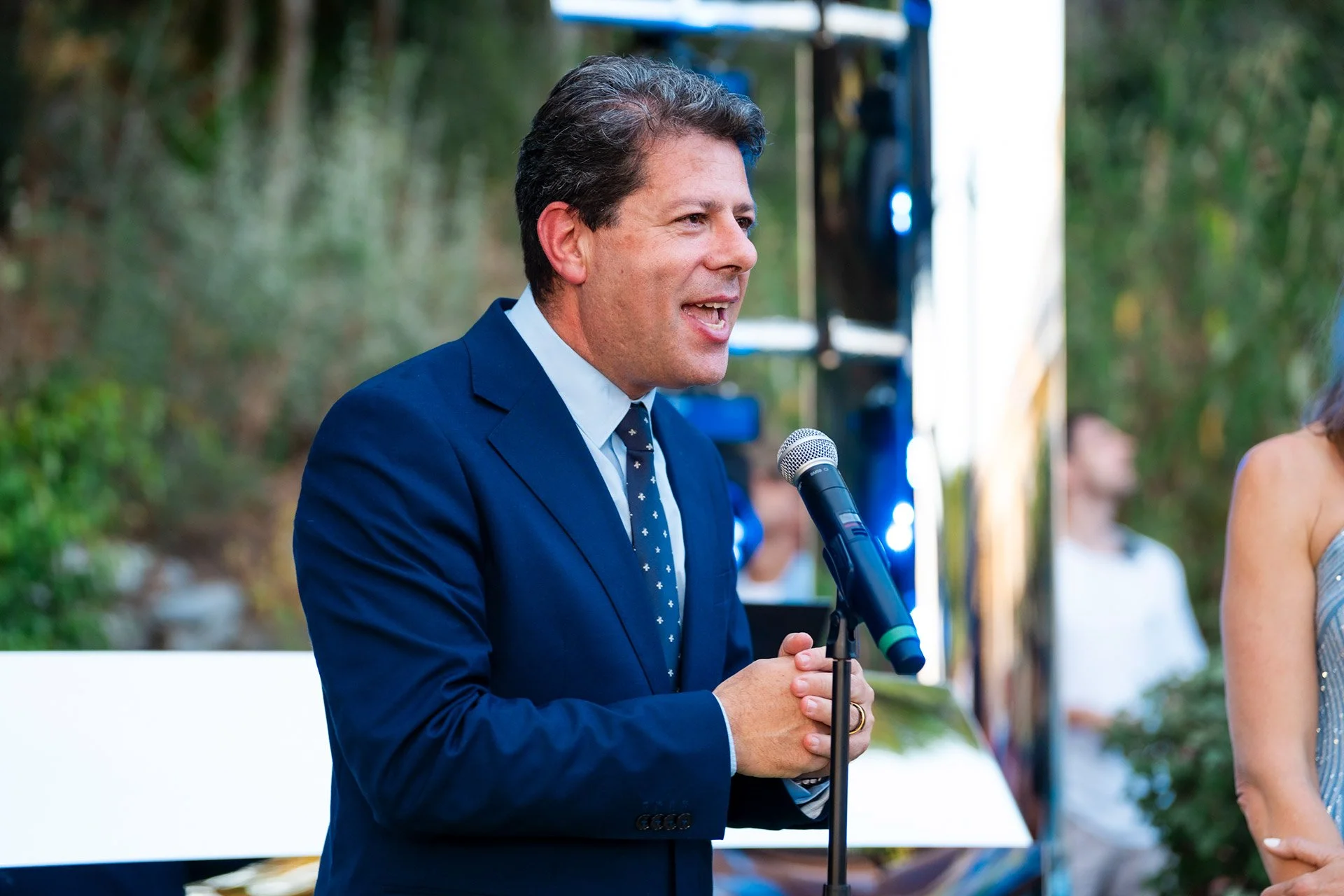 A man in a navy blue suit speaking into a microphone at an outdoor event, with people and greenery in the background.