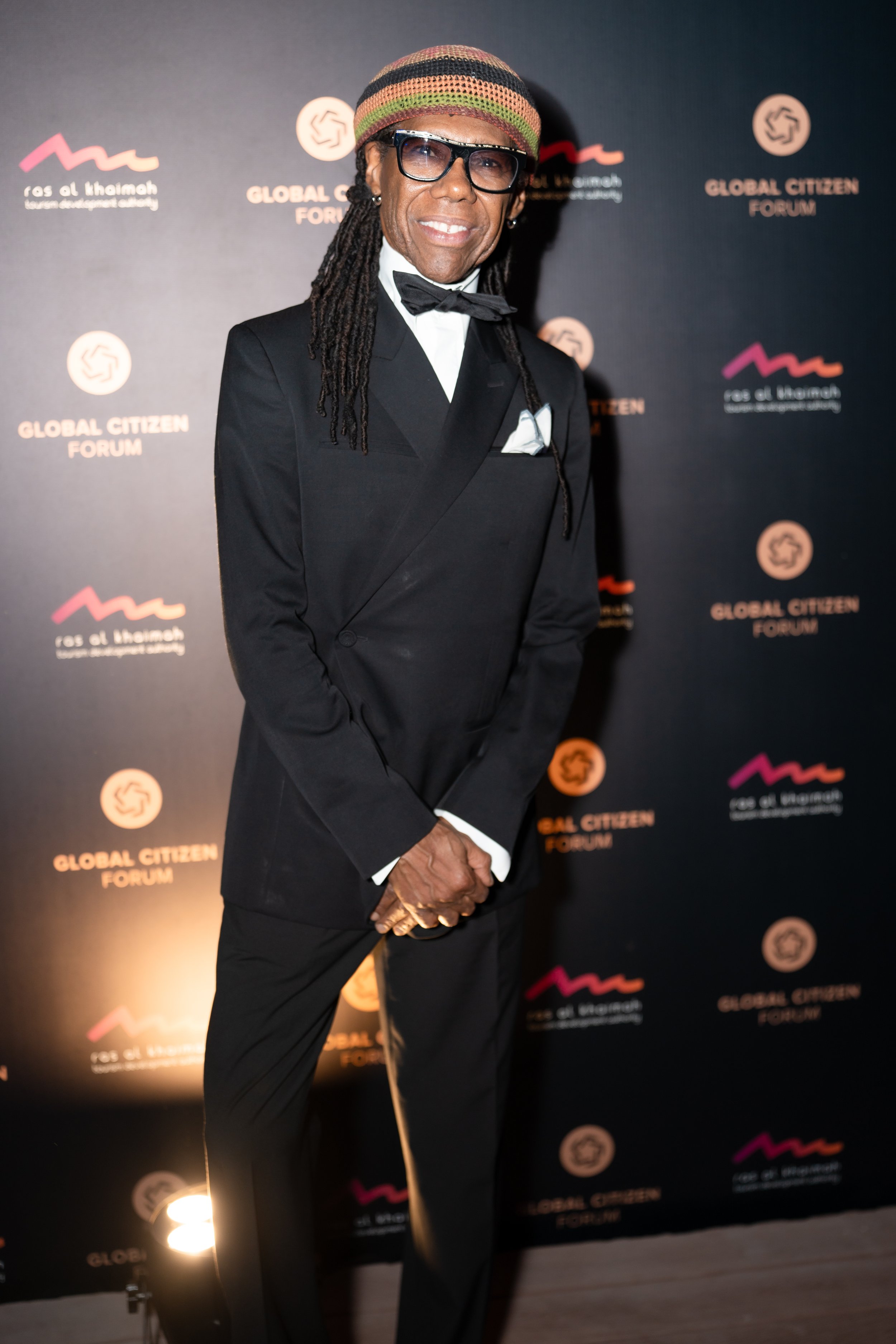 A man dressed in a tuxedo with a bow tie, wearing a multicolored hat and glasses, standing in front of a backdrop with the logo and name of the Global Citizen Forum.