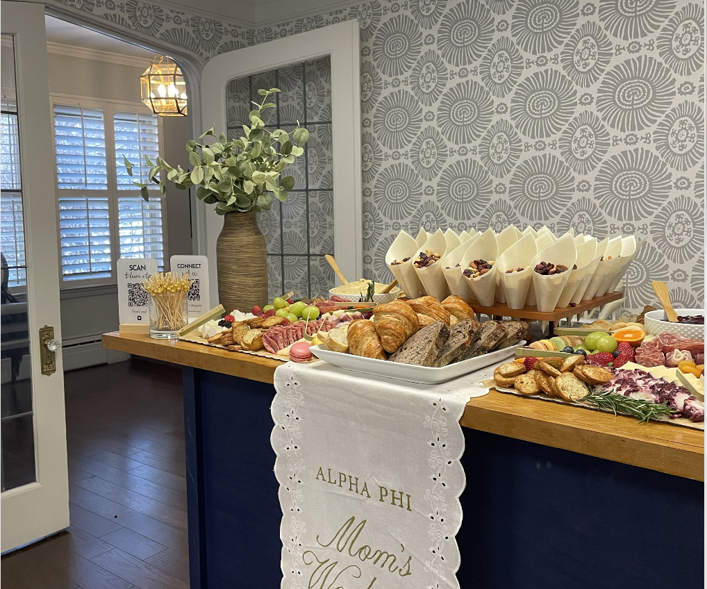 A brunch buffet table decorated with a white cloth embroidered with "Alpha Phi Mom's U" holding croissants, bread, cheese, vegetables, berries, grapes, charcuterie, parfaits, and a vase with green foliage, set in a room with patterned wallpaper and a