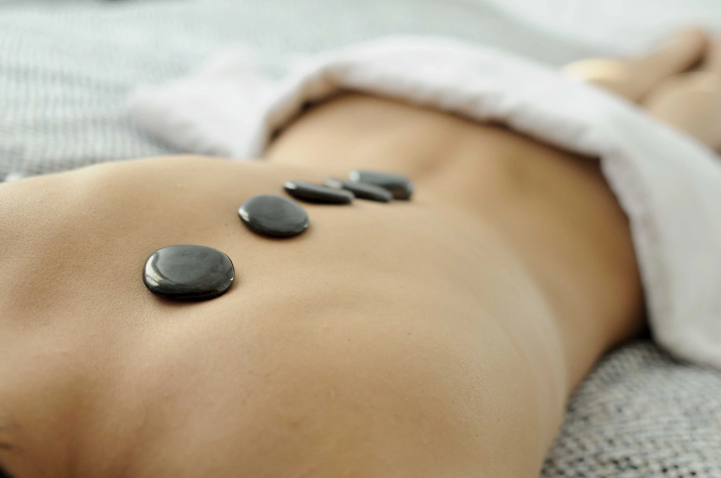 Close-up of a person's back with smooth skin, lying on a bed with hot stones placed along the spine, often used in hot stone therapy or massage.