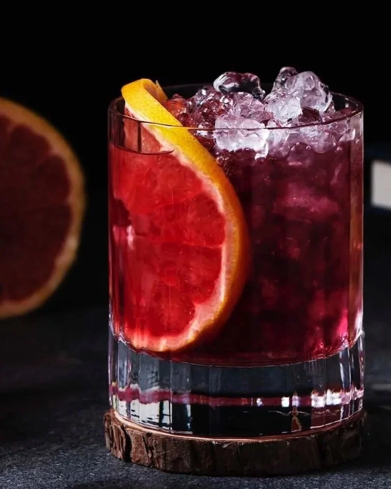 A glass of blood orange cocktail with ice and a slice of blood orange on the rim, surrounded by blood orange halves, a lemon, a lime, a cocktail jigger, a cocktail strainer, and stacked books against a dark background.