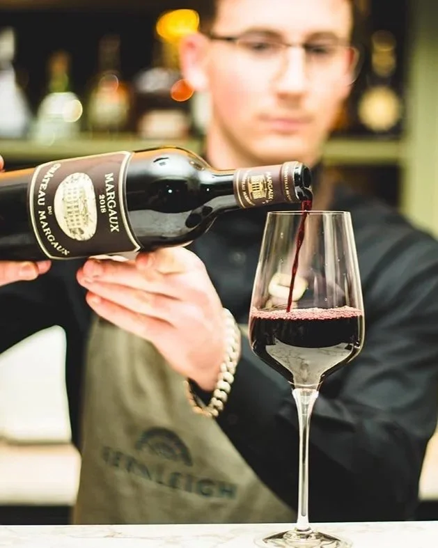 A bartender pours red wine from a bottle into a wine glass at a bar.