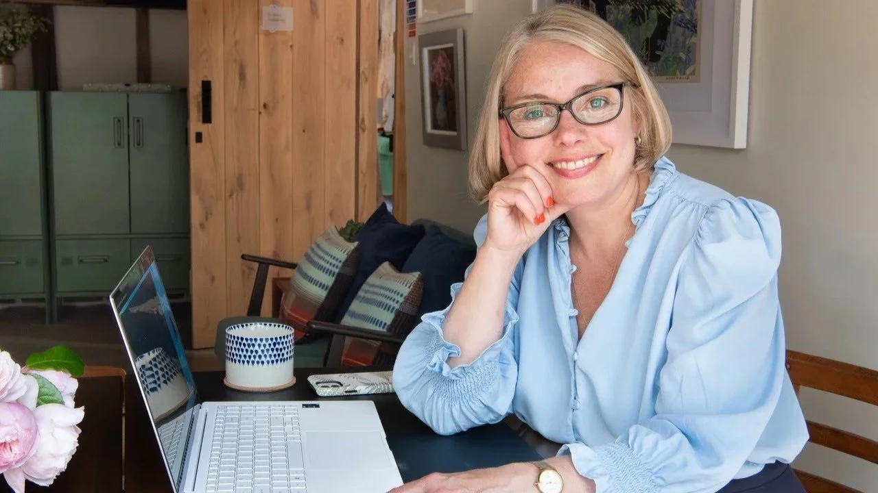 A woman with glasses and blonde hair sitting at a table, smiling and resting her chin on her hand with a laptop and decorative items in the background.