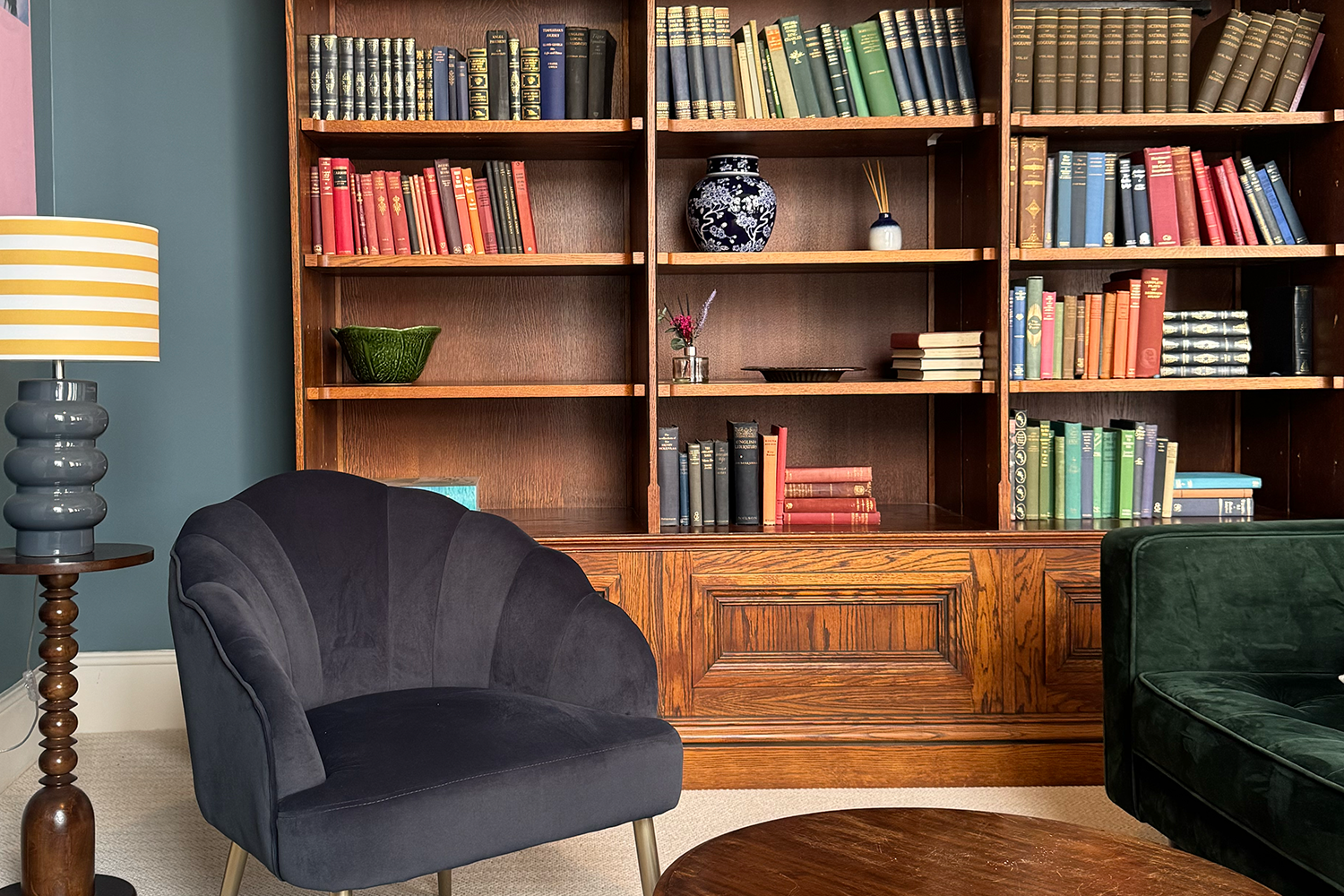 A room with a large wooden bookshelf filled with colorful books, a black velvet armchair, a green velvet armchair, a wooden coffee table, and a standing lamp with a striped shade on a base of stacked rounded shapes.