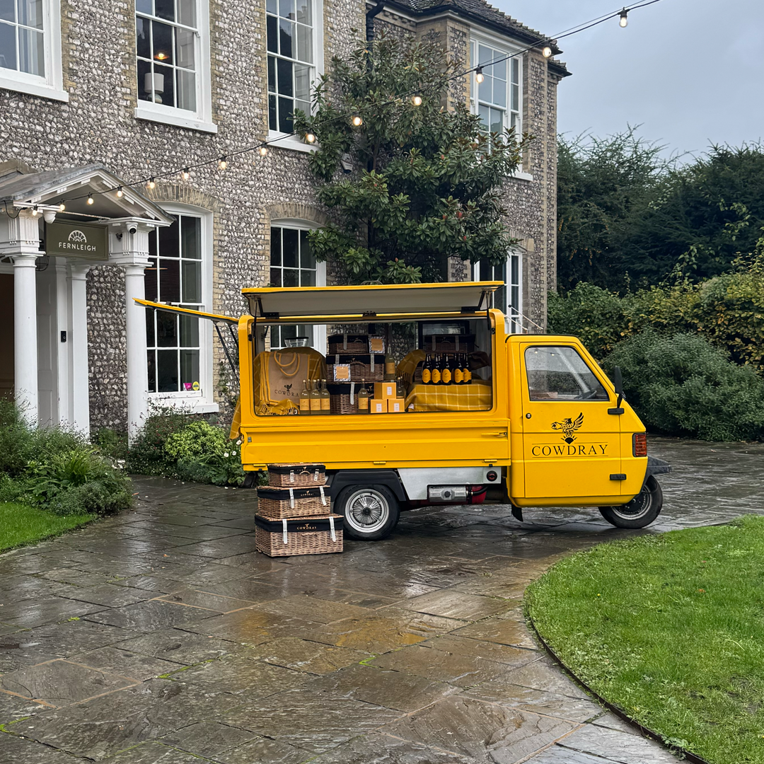 Yellow food truck with the name 'Cowdray' on the side, parked outside a large brick house on a wet stone driveway, with baskets and boxes of items in the open back and on the ground.