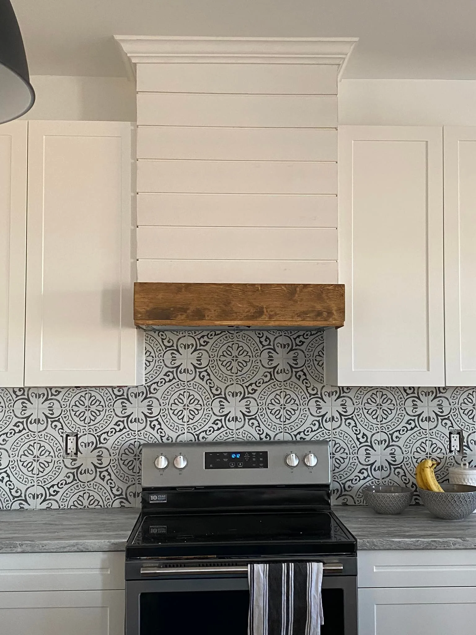 Kitchen with white cabinets, a black and stainless steel oven, decorative black and white tile backsplash, a wooden shelf above the stove, and bowls with bananas on the counter.