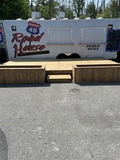 A food truck with the words "Road House" and the number "49" on it, with wooden planters and a small set of stairs in front, parked on a paved surface outdoors.