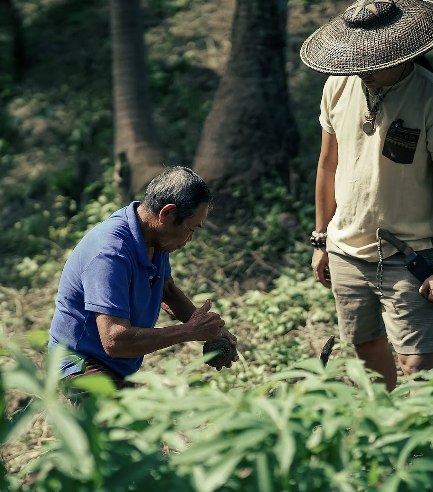 Two men in a lush, green forest, one kneeling and examining a small object or plant, the other standing and observing, both wearing casual outdoor clothing and accessories.