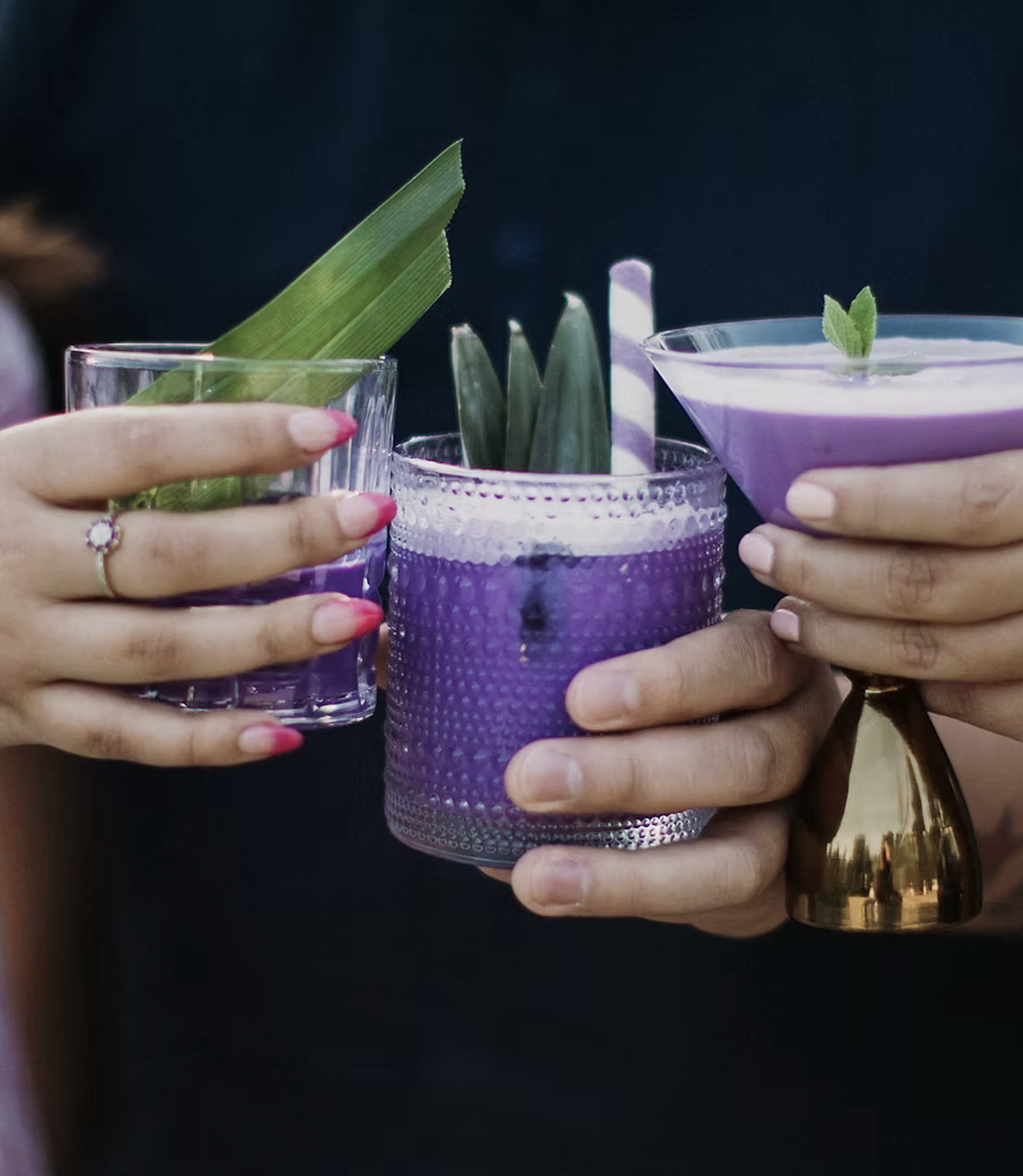 Three hands holding glasses of purple drinks garnished with green herbs and pineapple leaves, with colorful straws, against a dark background.
