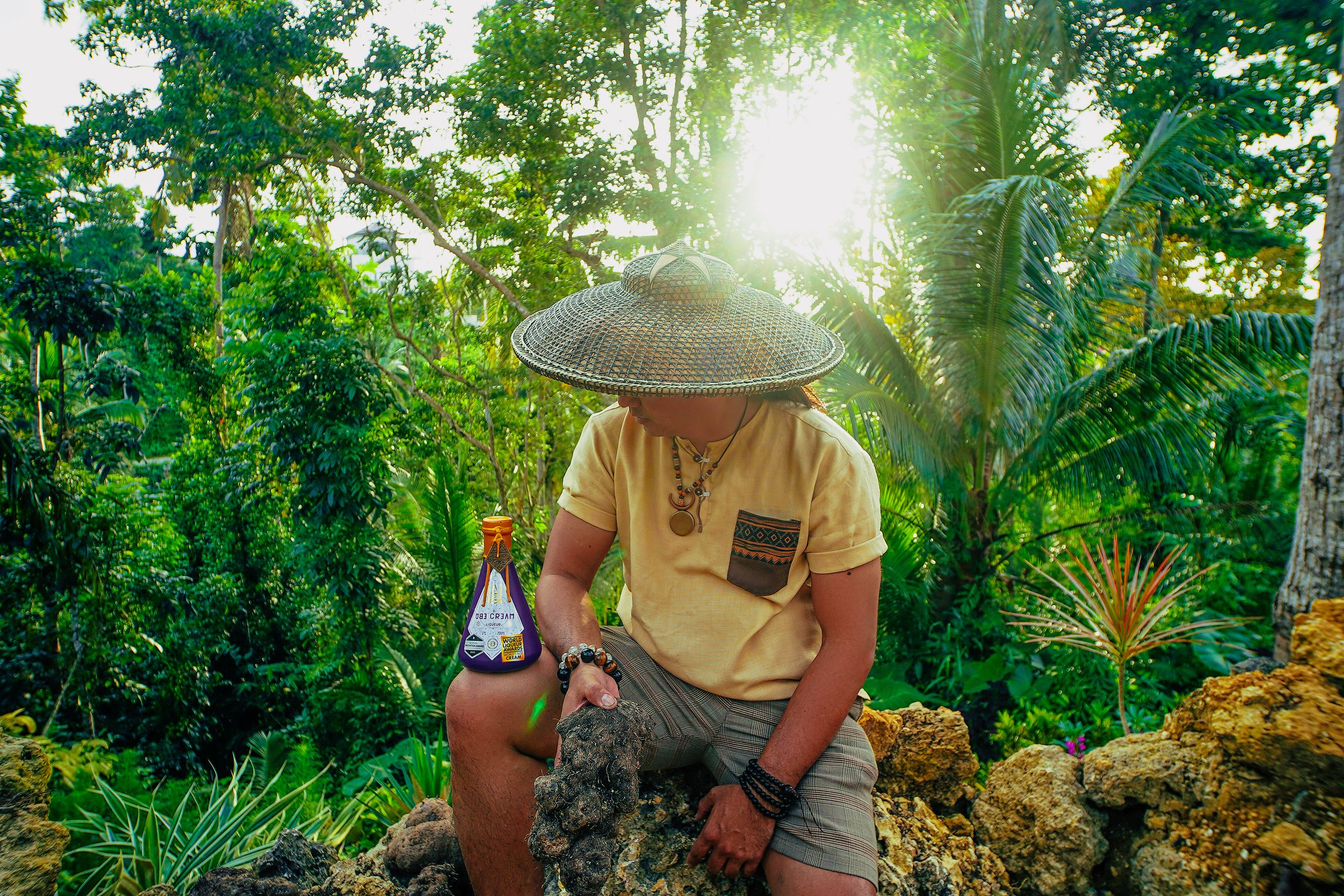 A person sitting outdoors on rocks in a lush jungle with big green leaves, wearing a wide-brimmed woven hat, a yellow shirt with patterned pocket, plaid shorts, and jewelry, holding a bottle of sauce.