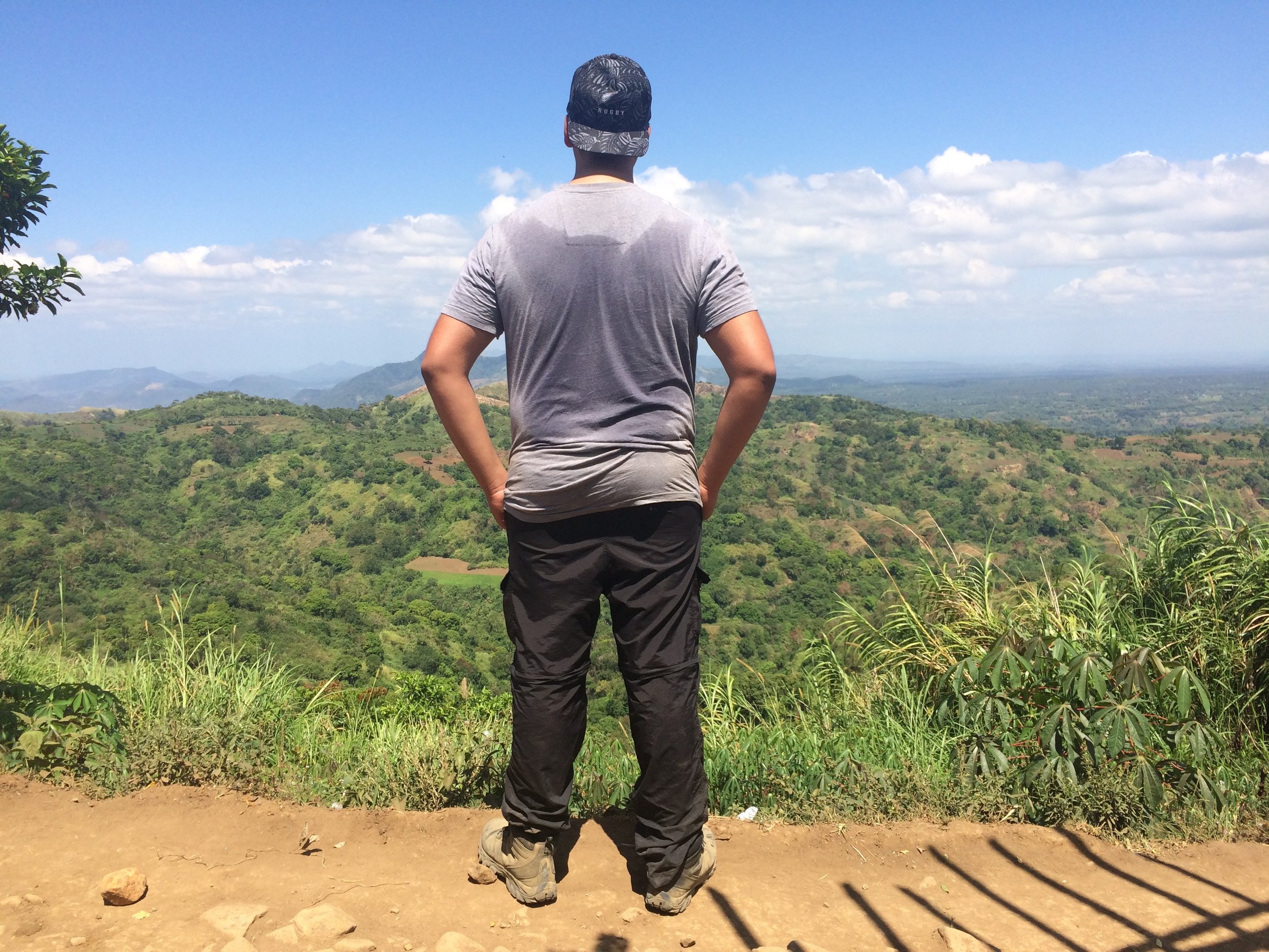 Man standing on a hilltop overlooking lush green mountains and a blue sky with clouds, facing away from the camera.