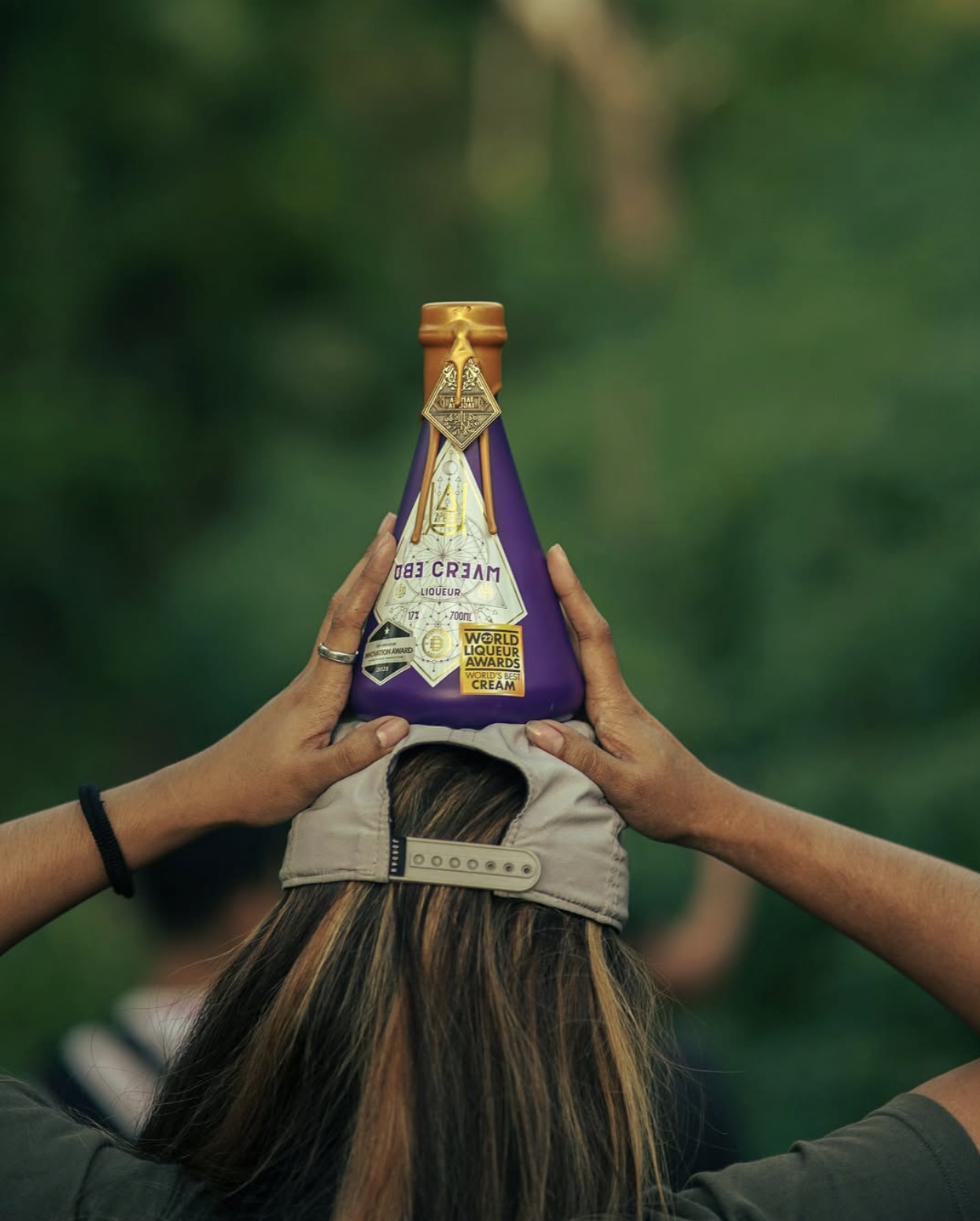 A person wearing a backward beige baseball cap, holding a purple bottle labeled "Cream Ed" above their head. The background is blurred, showing green outdoor scenery.