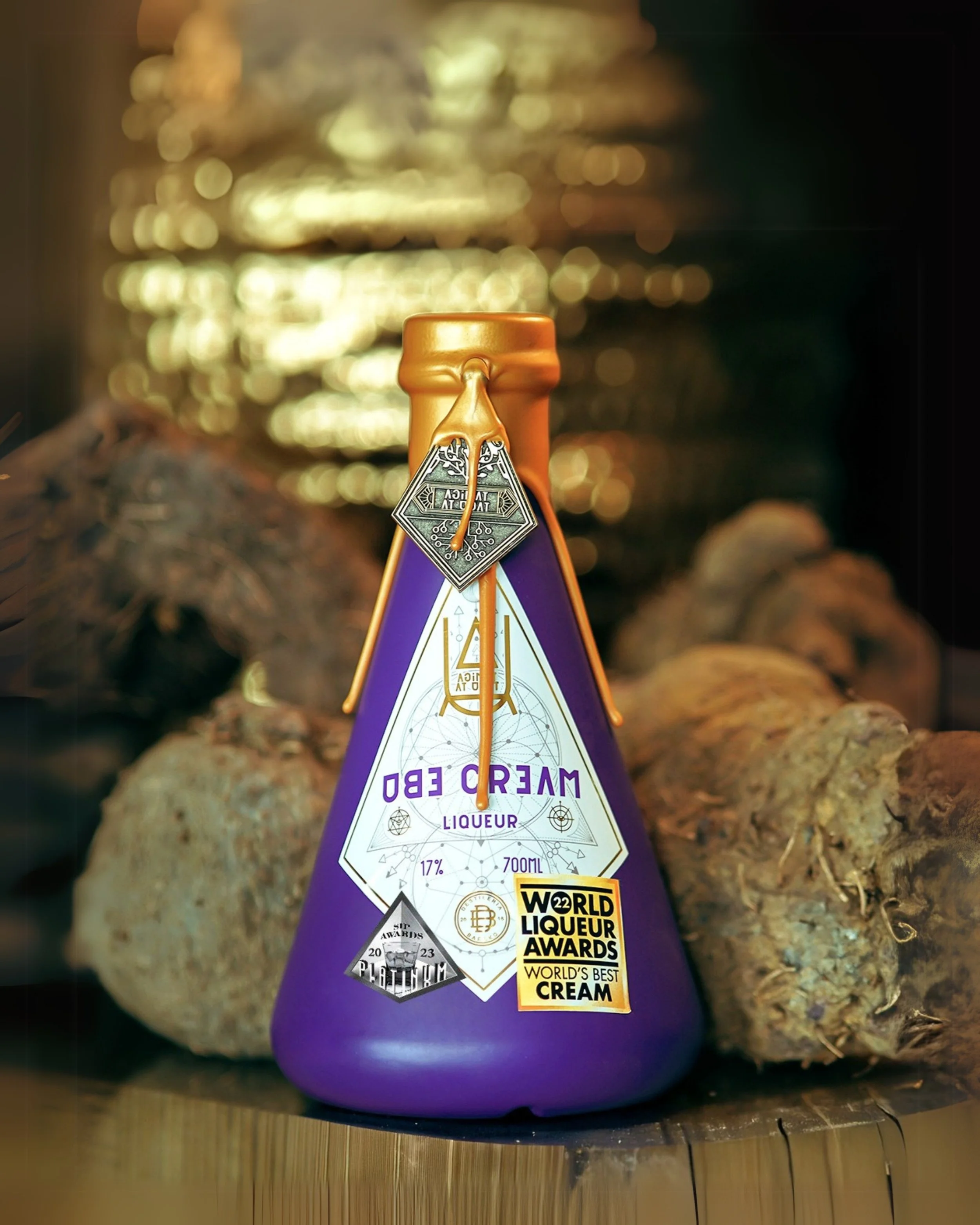 A purple and gold bottle labeled 'Cream Ed Liqueur' with awards stickers, sitting on a wooden surface with logs in the background.