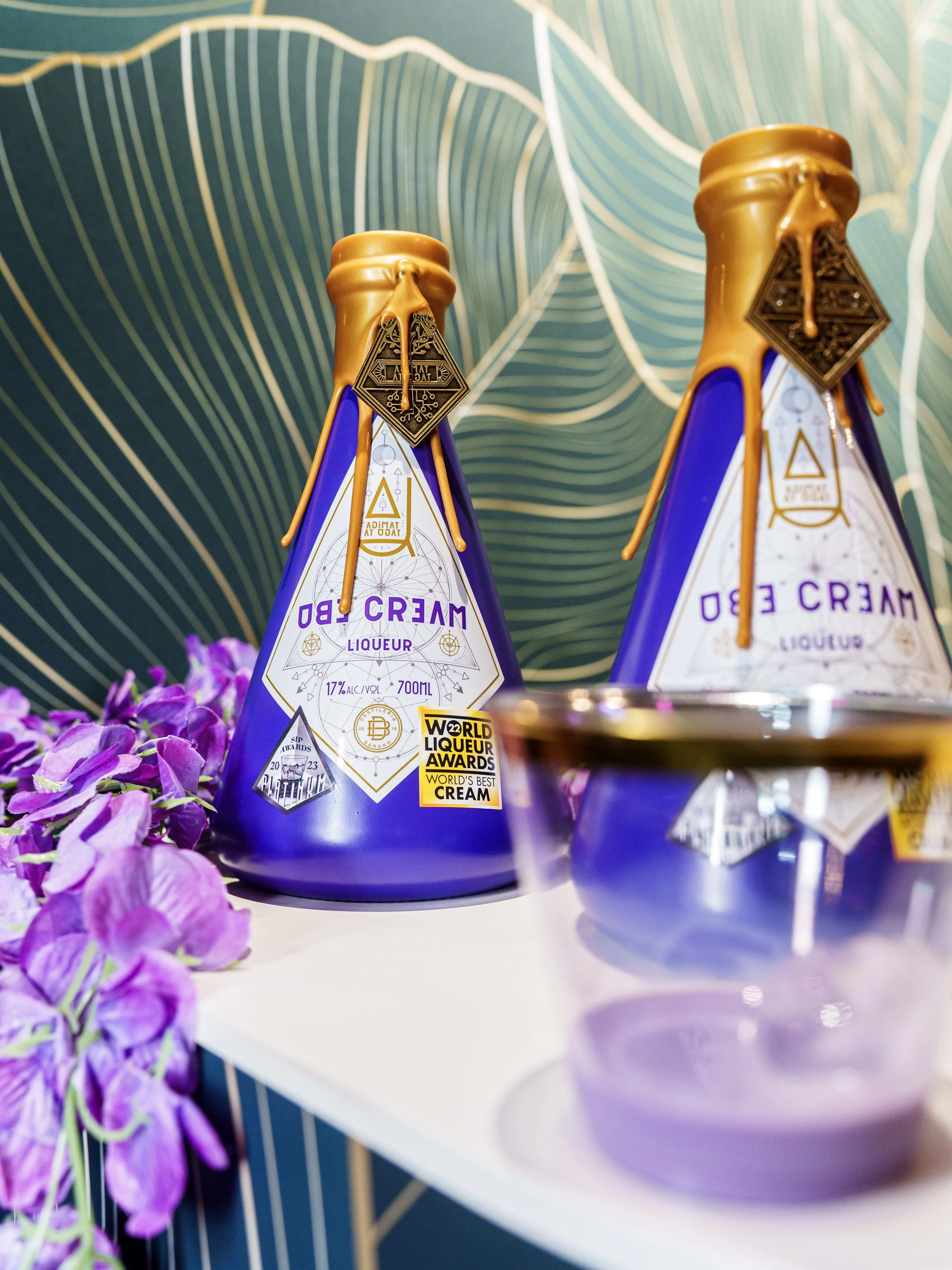 Two purple triangular bottles of whipped cream liqueur with gold caps and tags, placed on a white surface with purple flowers in the foreground and a teal decorated background.