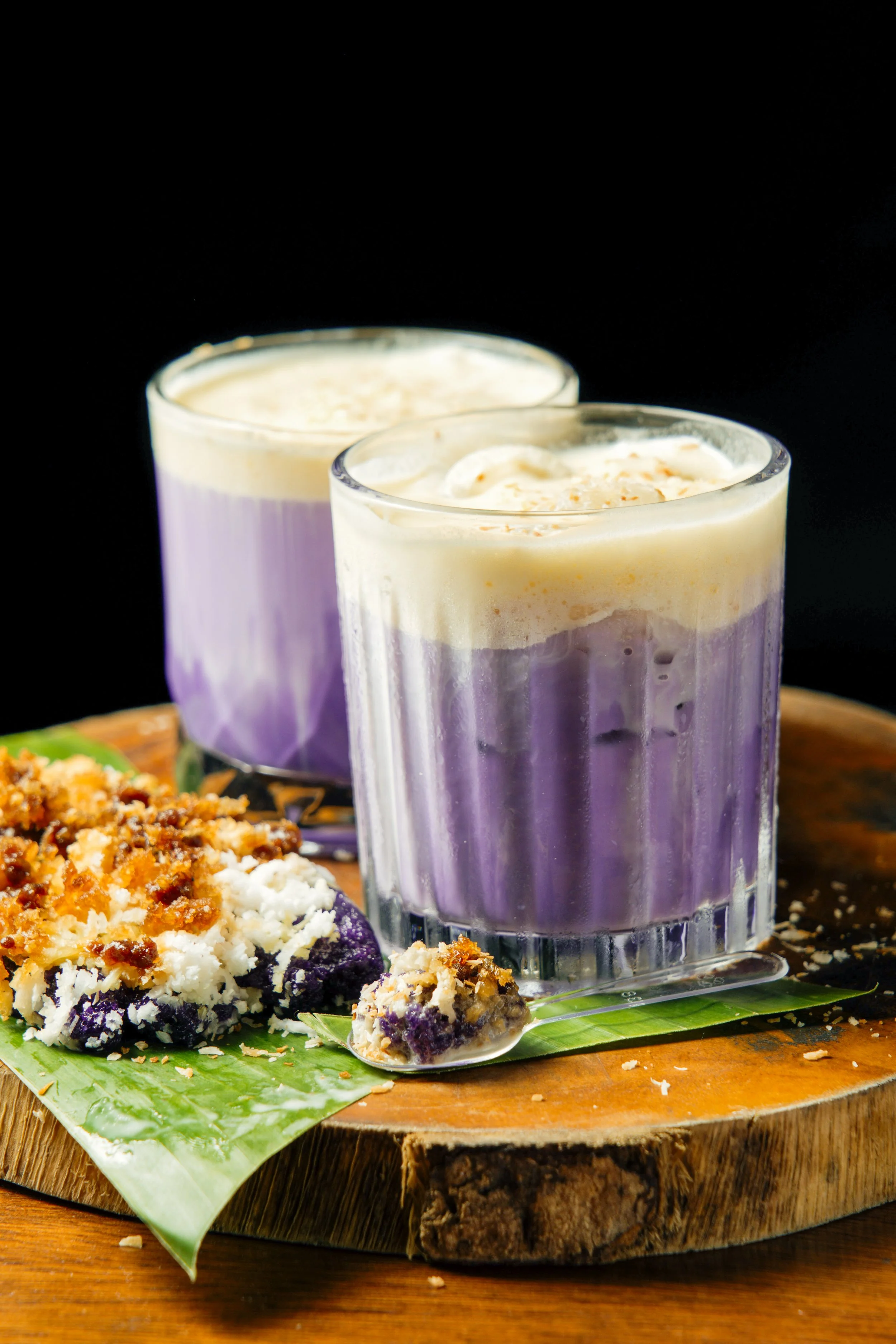 Two purple and white frosted drinks in glasses on a wooden serving board, with a slice of blueberry crumble topped with whipped cream on a green leaf, against a black background.