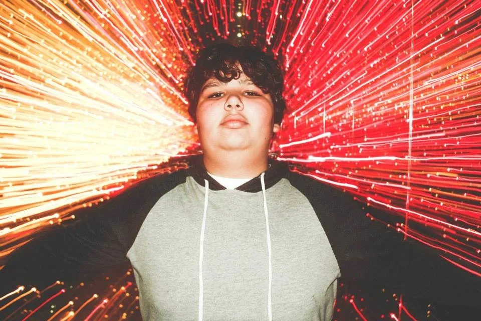 A young person with curly hair standing in front of a background with red and yellow streaks of light resembling fireworks or sparklers.