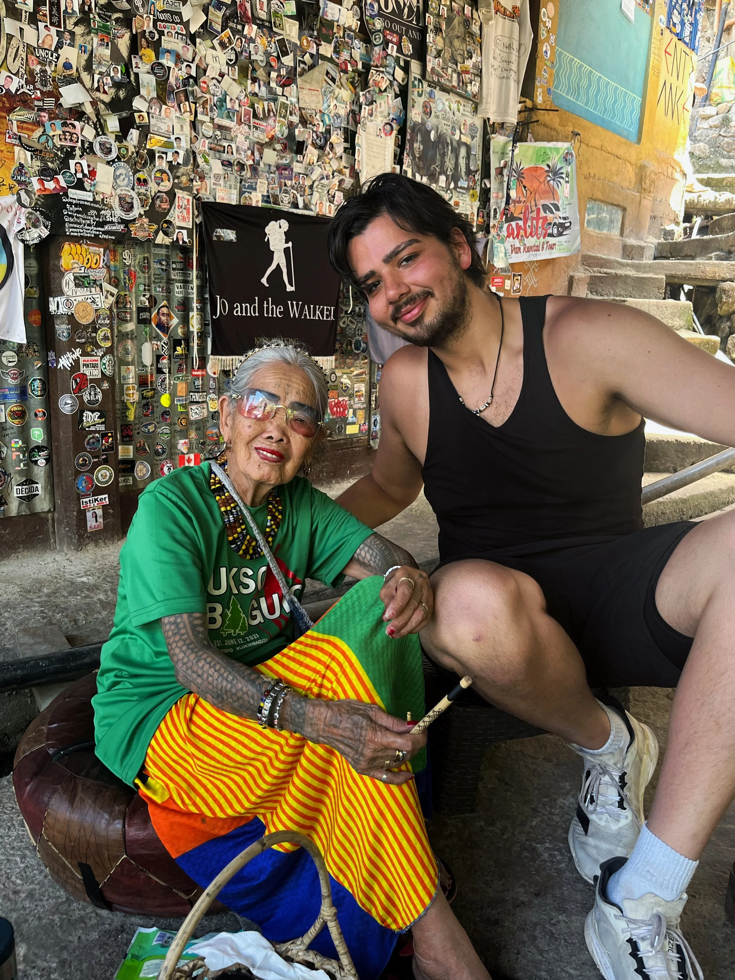 A young man and an elderly woman sitting together in front of a wall covered with stickers, photographs, and signs. The elderly woman wears sunglasses, colorful traditional clothing, and jewelry, holding a drumstick, while the young man wears a black tank top and shorts, smiling at the camera.