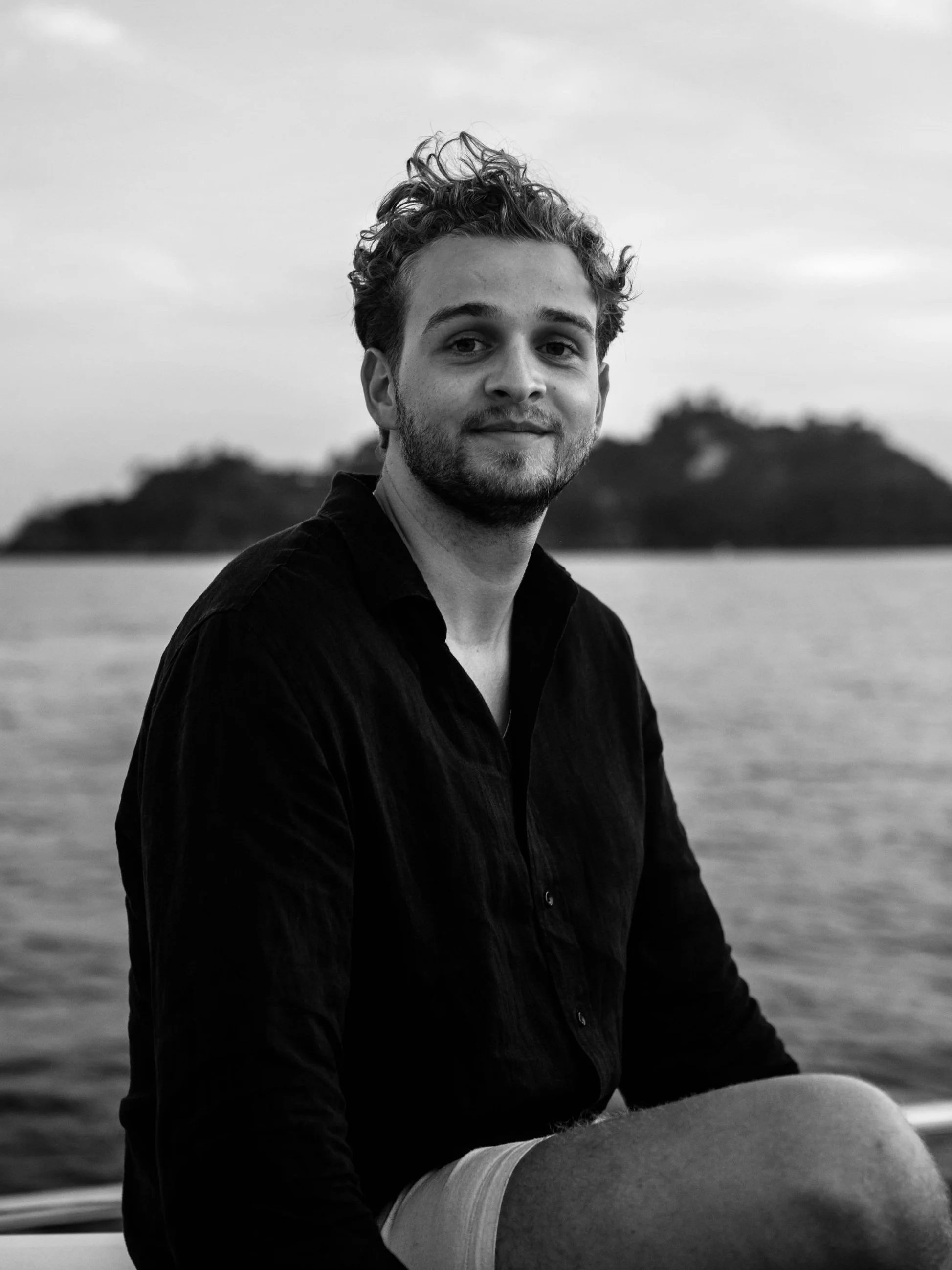 A young man with curly hair and a beard, wearing a dark shirt and light shorts, sitting outdoors near water with an island in the background, photographed in black and white.