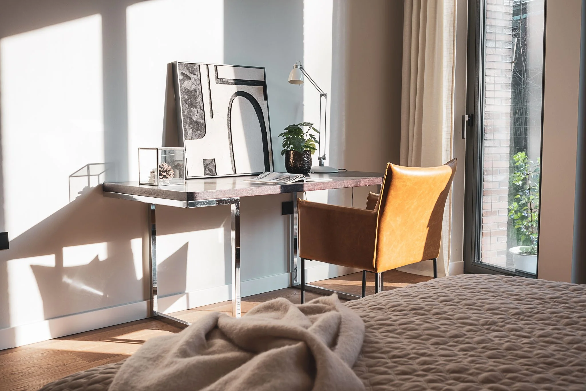 A bedroom with natural sunlight, featuring a bed with beige quilted bedding, a mustard-colored chair at a sleek desk with artwork, a potted plant, and a modern desk lamp, next to a glass door with curtains.