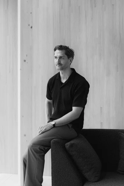 A man with short dark hair and a mustache sitting on a dark couch, wearing a black polo shirt and light-colored trousers, in a minimalist room with wooden walls.