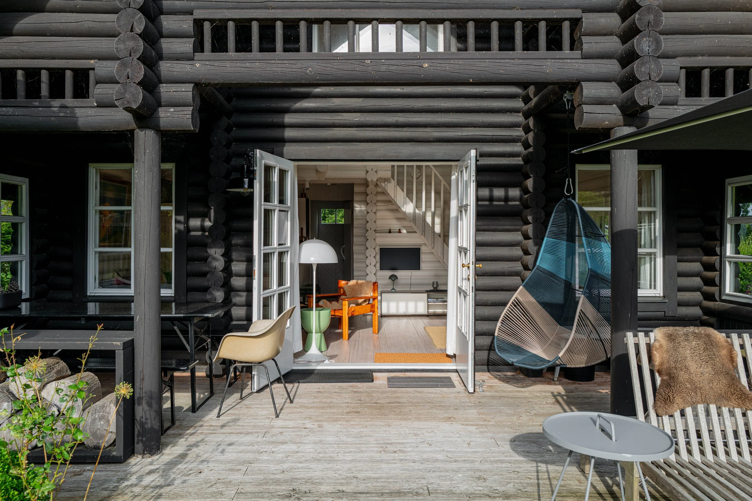 Cozy dining area in a wooden cabin with a rectangular wooden table, four chairs with cushions, bottles of alcohol on the table, large windows with white curtains, a beige rug, and a metallic pendant light hanging from the ceiling.
