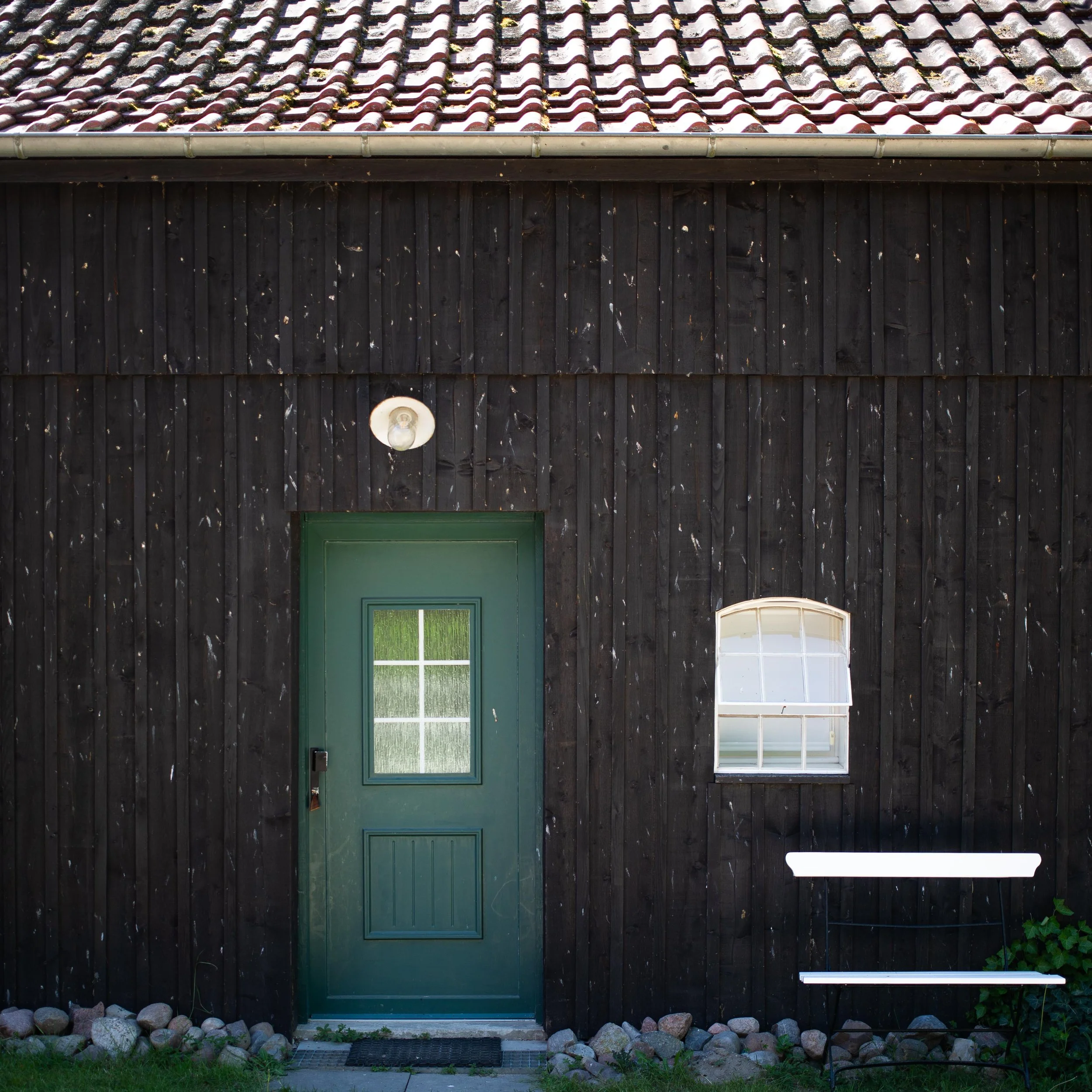 A rural scene viewed through a partially open dark wooden gate, with a grassy yard, a dirt path, a small chicken walking, and wooden and concrete buildings surrounded by green trees.