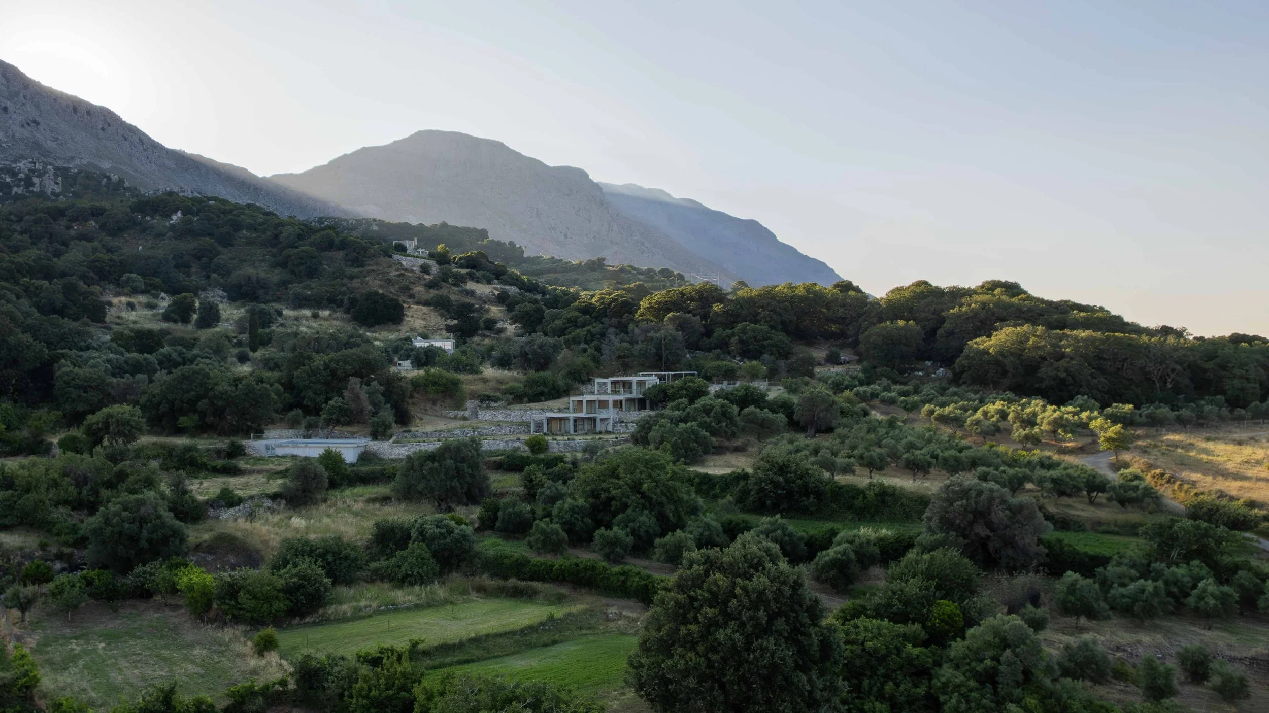 Hilly landscape with dense trees, a modern house, and mountains in the background