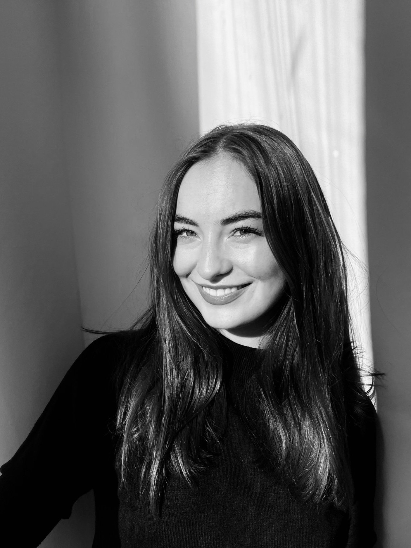 Black and white portrait of a young woman with long dark hair, smiling, wearing a black top, standing near a light-colored curtain.