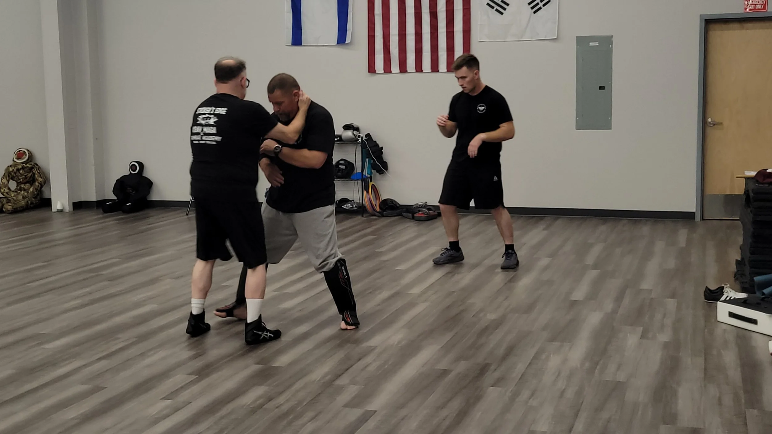 Three men training in a martial arts classroom, two engaged in a grappling drill, one standing nearby. Flags on the wall include the United States flag, South Korean flag, and a team flag. Equipment and gear are on the floor and wall-mounted shelves.